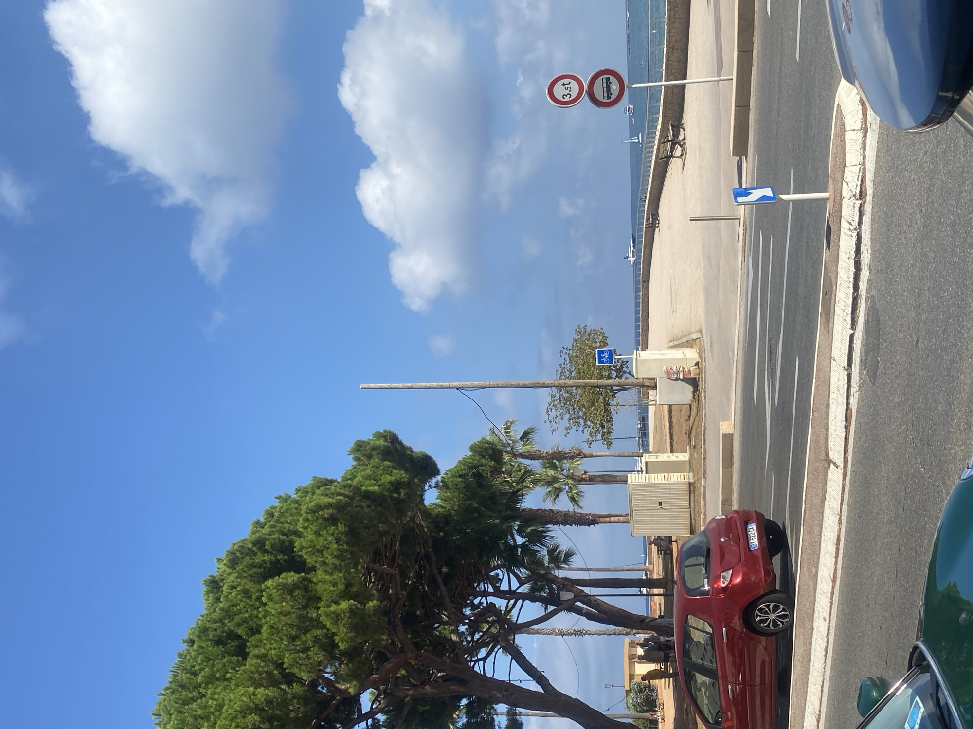 The image depicts a sunny day in Antibes, France, specifically near the Promenade du Port or a similar waterfront area. In the foreground, a red Renault Clio car is parked on the right side of a street, facing away from the camera. The car's license plate reads "BY 522 CX". A portion of a green car is visible in the bottom left corner.
The background shows a paved walkway with several benches, suggesting a public promenade. Beyond the promenade, the Mediterranean Sea is visible, with a few sailboats and a white airplane in the distance. Several palm trees and a large pine tree line the promenade, providing shade.
On the right side of the promenade, there are two traffic signs. The top sign is circular with a red border and indicates a weight limit of "3.5t". The bottom sign depicts a bus within a red circle with a diagonal red line, indicating that buses are prohibited. A blue and white directional sign pointing to the right is also present on the street. Several people are seen walking and standing near some light-colored structures on the left side of the promenade.