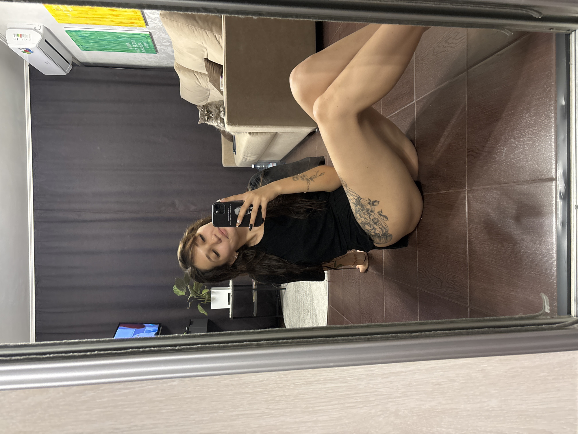 A young woman with long dark hair is sitting on the floor, taking a mirror selfie. She is wearing a black t-shirt and underwear, with tattoos on her legs and arms. In the background, a cat is sitting on a sofa, and there are paintings on the wall. The room appears to be a living room with a television and air conditioning unit. The image is taken in Novokuznetsk, Russia.