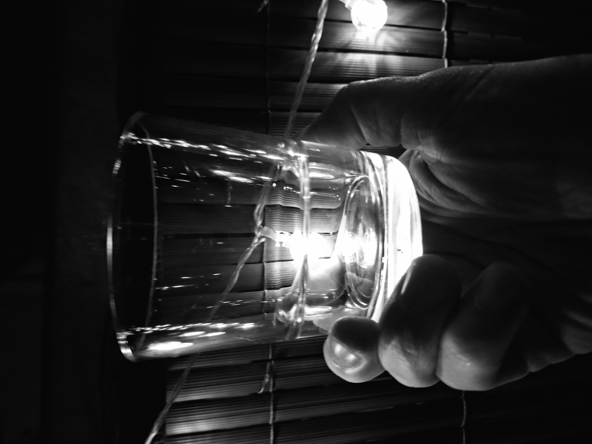 A close-up, black and white, low-angle shot shows a hand holding a clear glass filled with a liquid, likely water, indoors. The hand is gripping the glass from below. The glass has a string of small, round lights, possibly fairy lights, wrapped around it, and some of these lights are illuminated, casting a soft glow through the liquid. In the background, a textured, dark surface, possibly a bamboo mat or blinds, is visible, with more of the string lights scattered behind it. The lighting is dim, with a bright light source reflecting within the glass. The scene is devoid of any identifying landmarks or characteristics that would associate it with Sinzheim, Germany.