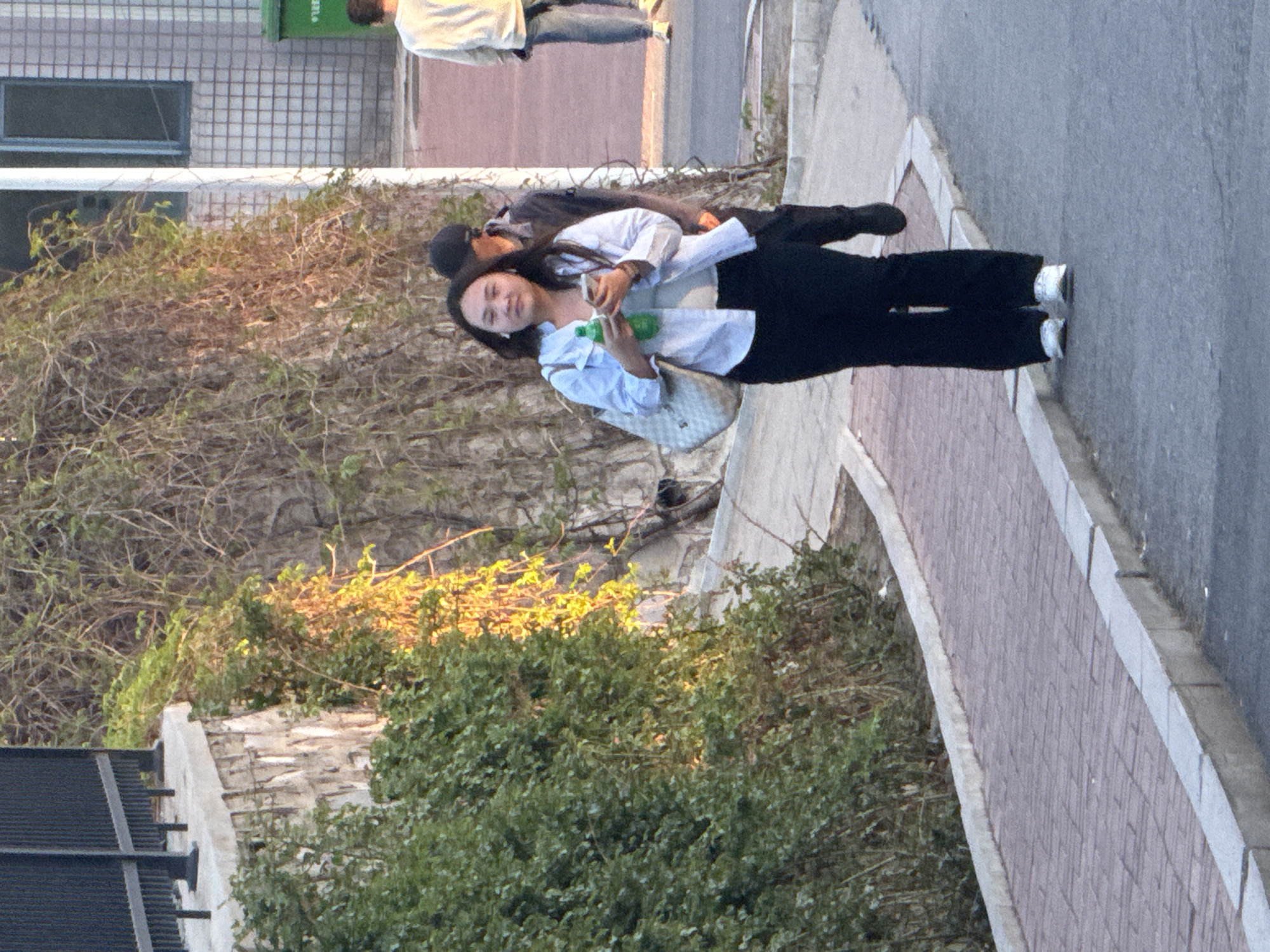 A woman is standing on a sidewalk in Zhongtun, China. She has long dark hair and is wearing a light blue shirt, black pants, and silver sneakers. She is holding a green water bottle and a cell phone. She has a white handbag on her shoulder. Another person is visible behind the woman, wearing a black cap and a black jacket.
The sidewalk is paved with red brick and gray concrete. There is a bush and some dead branches behind the woman. In the upper left corner of the image is a building. In the upper part of the image, two people are visible from the waist down. The image is taken from slightly above.
