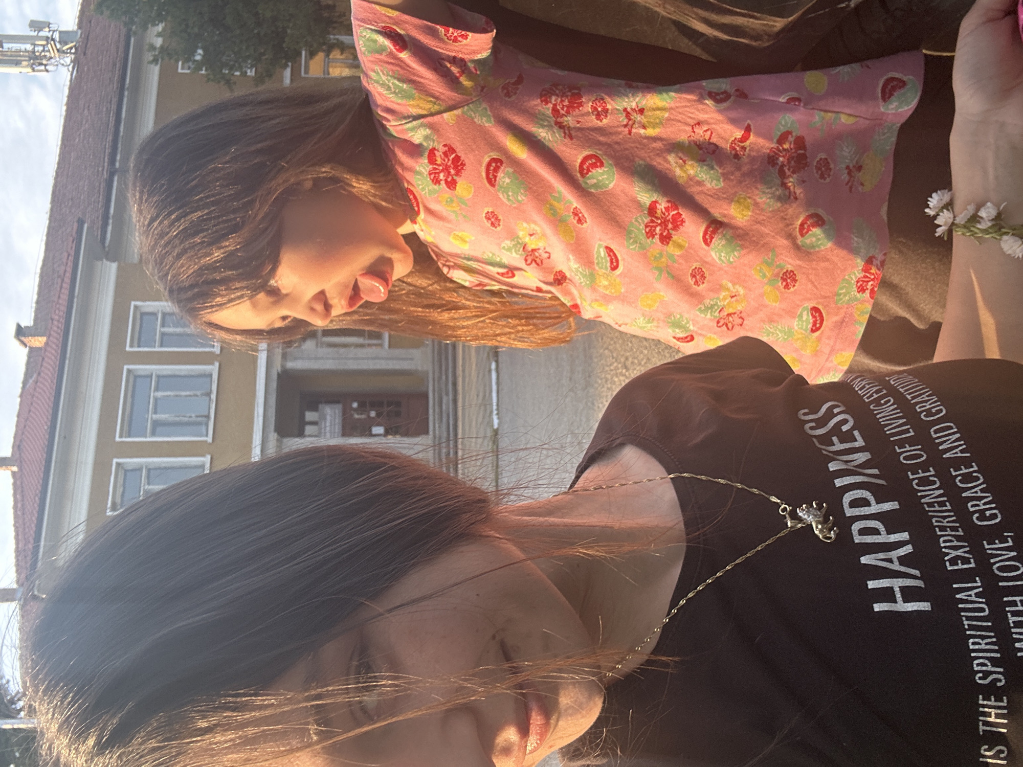 Two females, a girl and an adult, are positioned close to each other outdoors. The girl is wearing a pink patterned shirt, and the adult is wearing a dark t-shirt with the word "HAPPINESS" printed on it and a necklace with a pendant. The girl has her tongue out. The adult has a bracelet made of white flowers on their wrist. Behind them is a building with a red roof and multiple windows, appearing to be a public building with a central door. The location is Resen, Bulgaria.