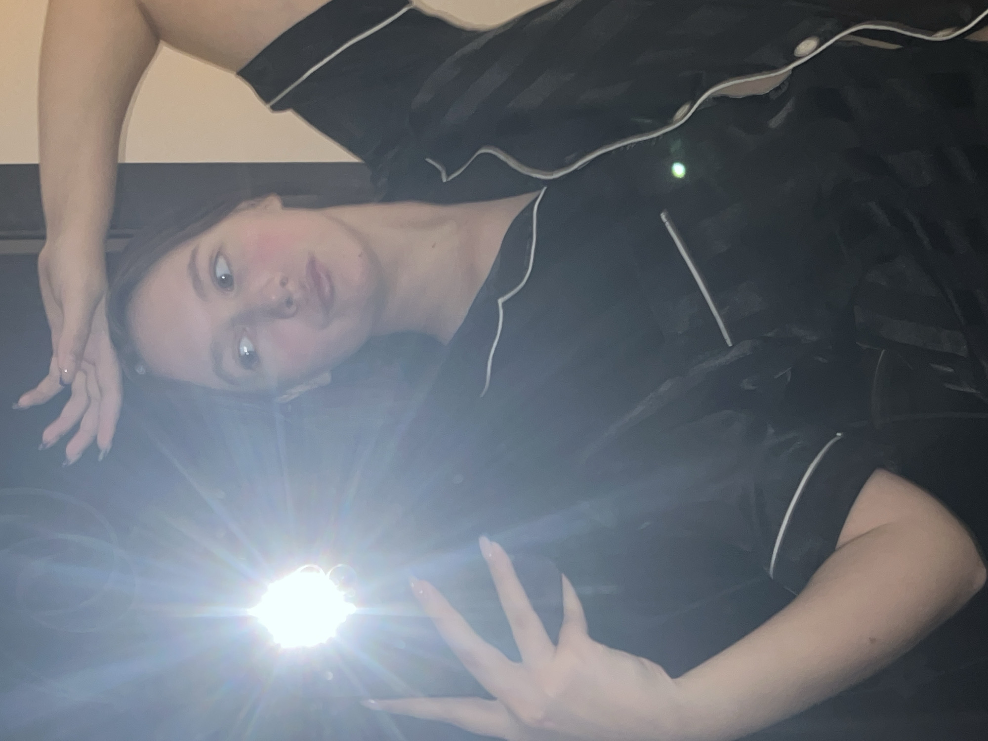 The image shows a person with pale skin and shoulder-length dark hair. They are wearing black pajamas with white piping. They are lying down. One hand is above their head and the other is near a bright light that is partially obscuring the lower portion of the image. The background is indistinct, but appears to be a black surface against a light-colored wall. The location is not discernible from the image.