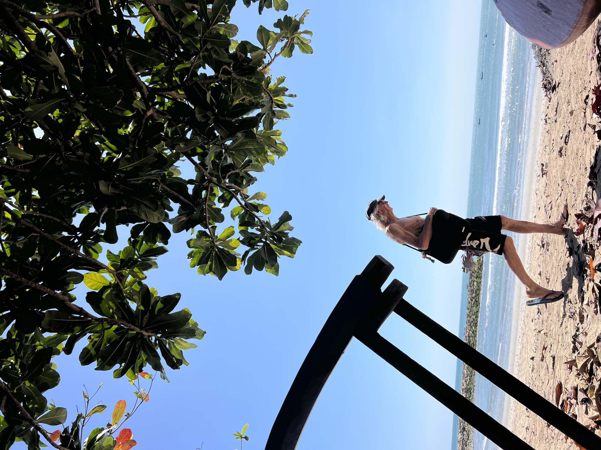The image depicts a man walking on a beach in Kuta, Indonesia. He is wearing a cap, glasses, and swim trunks, with a sling bag over his shoulder. The man is in motion, with his legs slightly apart as if mid-stride. He is walking away from the viewer, towards the ocean. In the background, the turquoise waters of the Indian Ocean meet a clear blue sky, with a few boats visible on the horizon. The beach itself is sandy, with scattered leaves and a large, dark, abstract structure in the foreground. Lush green foliage from a tree hangs over the scene from the top, casting shadows on the beach. The overall atmosphere is serene and tropical.