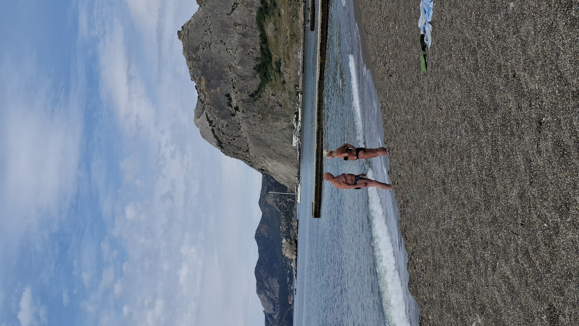 The image, currently oriented sideways (rotated 90 degrees counter-clockwise), depicts a coastal scene featuring a pebble beach, the sea, and dramatic mountainous terrain under a partly cloudy sky.
In the midground, two adults, an older man and woman, stand at the water's edge on the pebble beach. The woman, to the left (when correctly oriented), wears a dark two-piece swimsuit and has blonde hair tied back. The man, to her right, is bald and wears patterned swim trunks. They both face towards the water, which features gentle waves breaking on the shore. On the far right of the beach, a blue and a green item of clothing or towel are laid out.
The background is dominated by a large, steep, rocky mountain on the left side, with some sparse green vegetation. Further out in the calm sea, a concrete pier or breakwater extends, and a small white sailboat is visible. In the distant background, more dark, rugged mountains line the horizon. The sky is blue with scattered, soft white clouds, suggesting a clear day, likely late morning or early afternoon, given the bright, even lighting. There is no visible text or discernible emotional expression from the individuals.
