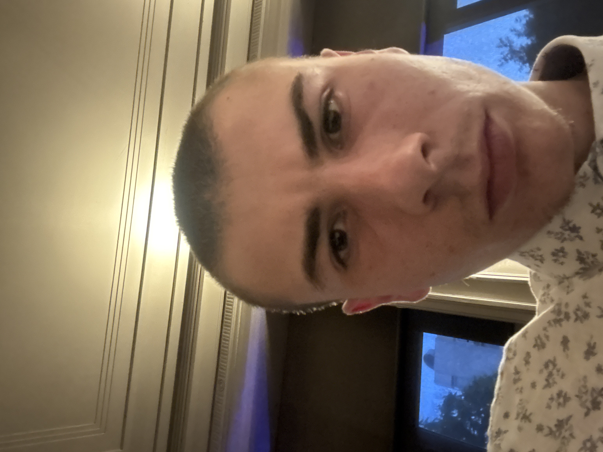 The image is a close-up selfie of a person with a shaved head, facing the camera at an angle. The person has light skin, dark eyebrows, and brown eyes. They are wearing a white shirt with a floral pattern. The background includes a wall with vertical decorative moldings. A window is visible, showing a blurry scene with trees and a building exterior, suggesting an outdoor environment. The image does not provide enough information to determine the precise location in Plovdiv, Bulgaria.