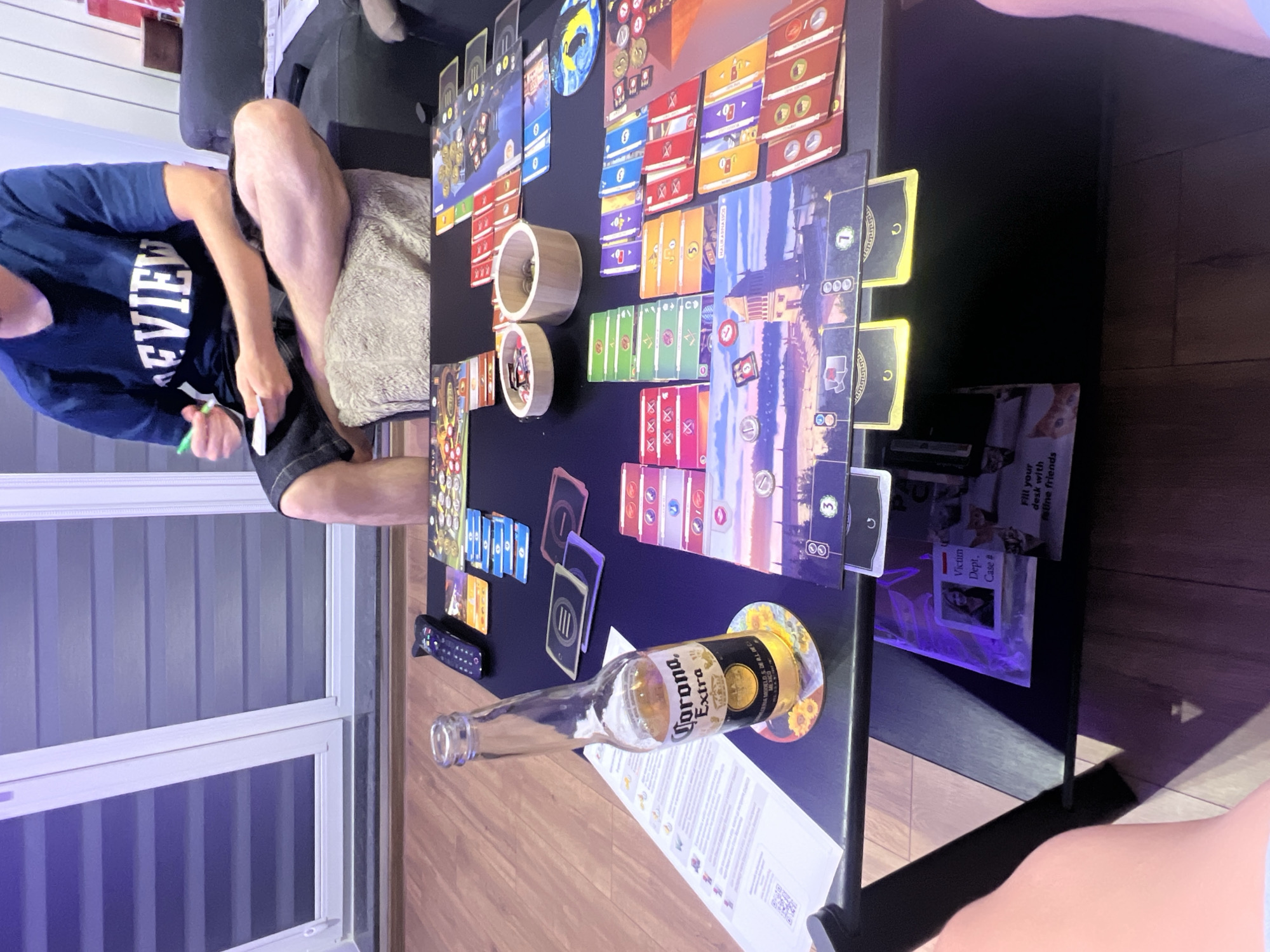 The image shows a person playing a board game at a coffee table. The person is wearing a dark blue t-shirt with the word "REVIEW" printed on it in white letters, and dark shorts. They are holding a green marker and a piece of paper, seemingly making notes. The coffee table is covered with various board game components, including cards, tokens, and dice. A bottle of Corona Extra beer is prominently displayed on a coaster with a sunflower design. There are also two wooden bowls filled with tokens. In the background, a couch and a window with horizontal blinds are visible. The floor is wooden. A remote control is also on the table. The scene is indoors. There is no discernible information that suggests the location is in Sofia, Bulgaria.