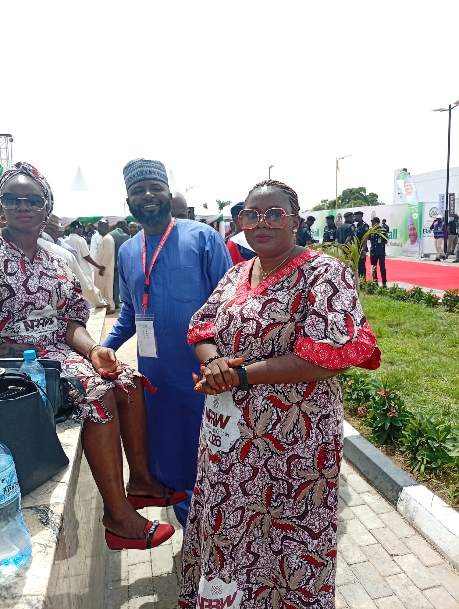 This outdoor photograph captures three individuals posing at an event in Unguwan Sanusi, Nigeria, under bright daylight. In the foreground, a man wearing a blue kaftan, a traditional patterned cap, and a beard smiles broadly. He has an ID badge around his neck which prominently features the Chinese characters "工作证" (Work Permit), alongside other text like "APE CROC CITY 200" and "NAFD."
He is flanked by two women dressed in matching patterned outfits featuring red, brown, and white designs. To his left, a woman is seated on a low concrete wall or bench, wearing a headwrap, dark sunglasses, and red flat shoes. A "Faya" brand water bottle and a black handbag are visible beside her. To the man's right, another woman stands, wearing red-rimmed glasses and an ID badge that reads "NRW 226 State Chapter" and "Kalimahanter."
The background indicates a large outdoor event, possibly a conference or festival. Tents and various banners are visible, with text including "Hall," "Kaduna," and "ENBRIDGE." A red carpet stretches into the distance, and uniformed personnel along with other attendees can be seen further back. The bright, clear sky suggests it is daytime. The scene is set on paved ground with patches of green foliage, contributing to a vibrant and communal atmosphere.