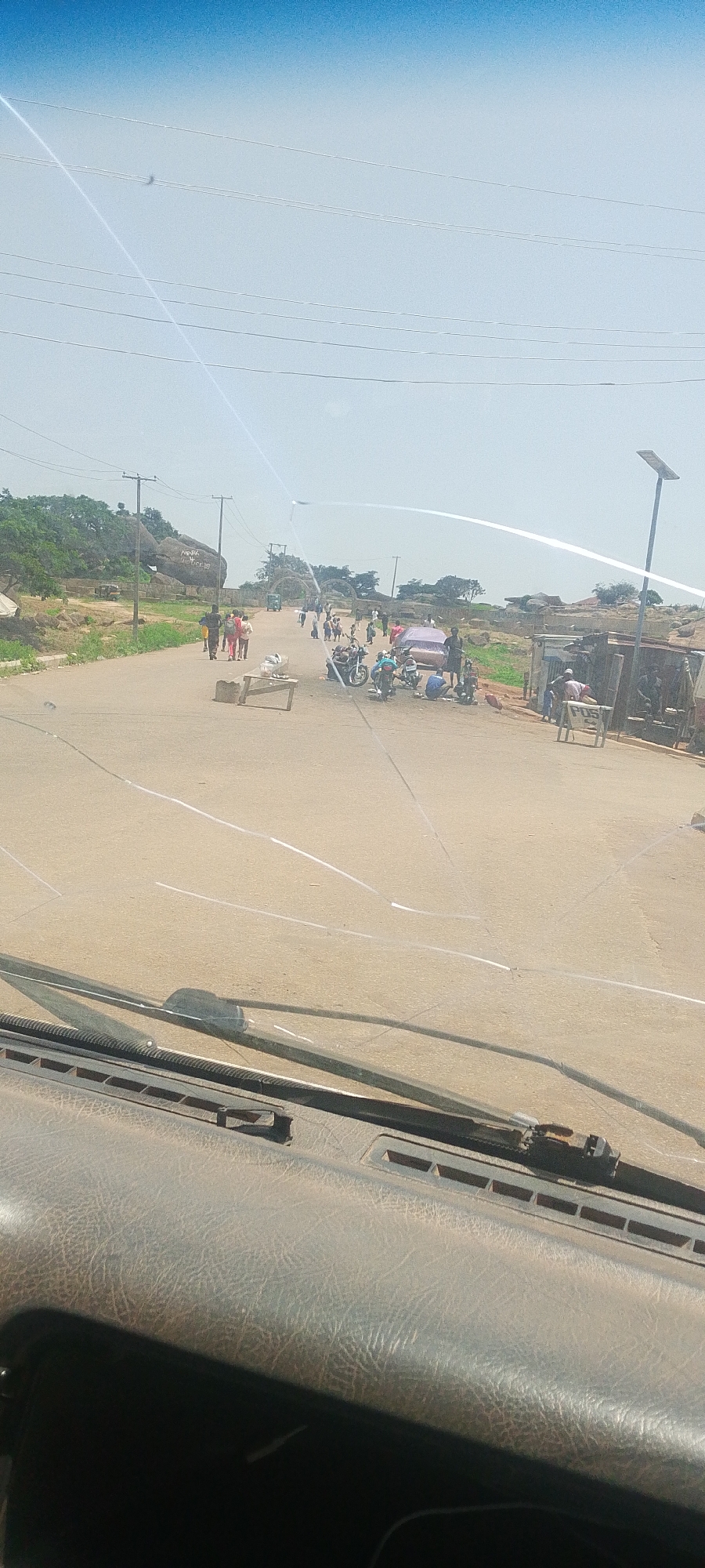 The image depicts a busy street scene in Jos, Nigeria. In the foreground, a car's cracked windshield is visible, obscuring parts of the view. The road itself is paved and appears to be a main thoroughfare, with several people and vehicles gathered.
On the left side of the road, a group of individuals, some carrying bags, are walking. A few motorcycles are parked on the right side of the road, where a small stall or shop with a POS (Point of Sale) sign is also present. A covered object, possibly a vehicle or cargo, is situated near the motorcycles.
Further down the road, several people are seen interacting. There's a small, possibly mobile, cart on the road. In the background, there are rocky outcrops and scattered vegetation, characteristic of Jos's landscape. Power lines and poles run along the street, and a solar-powered street light is visible. The overall atmosphere suggests a lively market or gathering place.