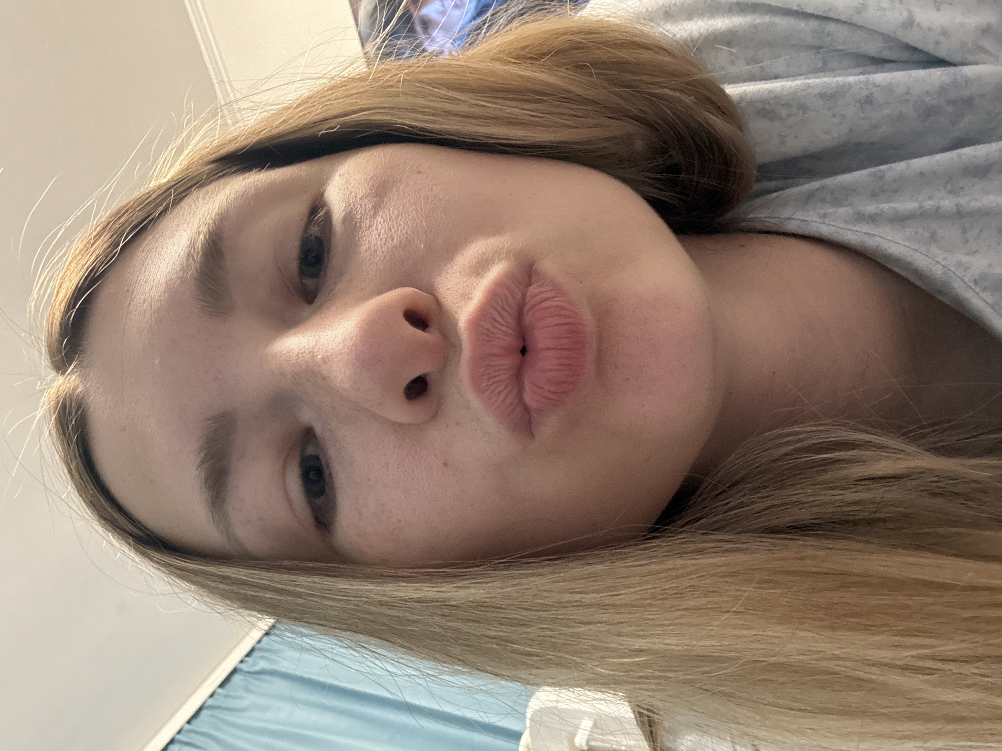 A close-up, angled selfie of a young woman with fair skin, blonde hair, and dark eyes. Her lips are pursed in a kissing pose. The background shows a light blue fabric and a white surface, possibly a bed and wall. No indication that the picture was taken in Moscow, Russia.