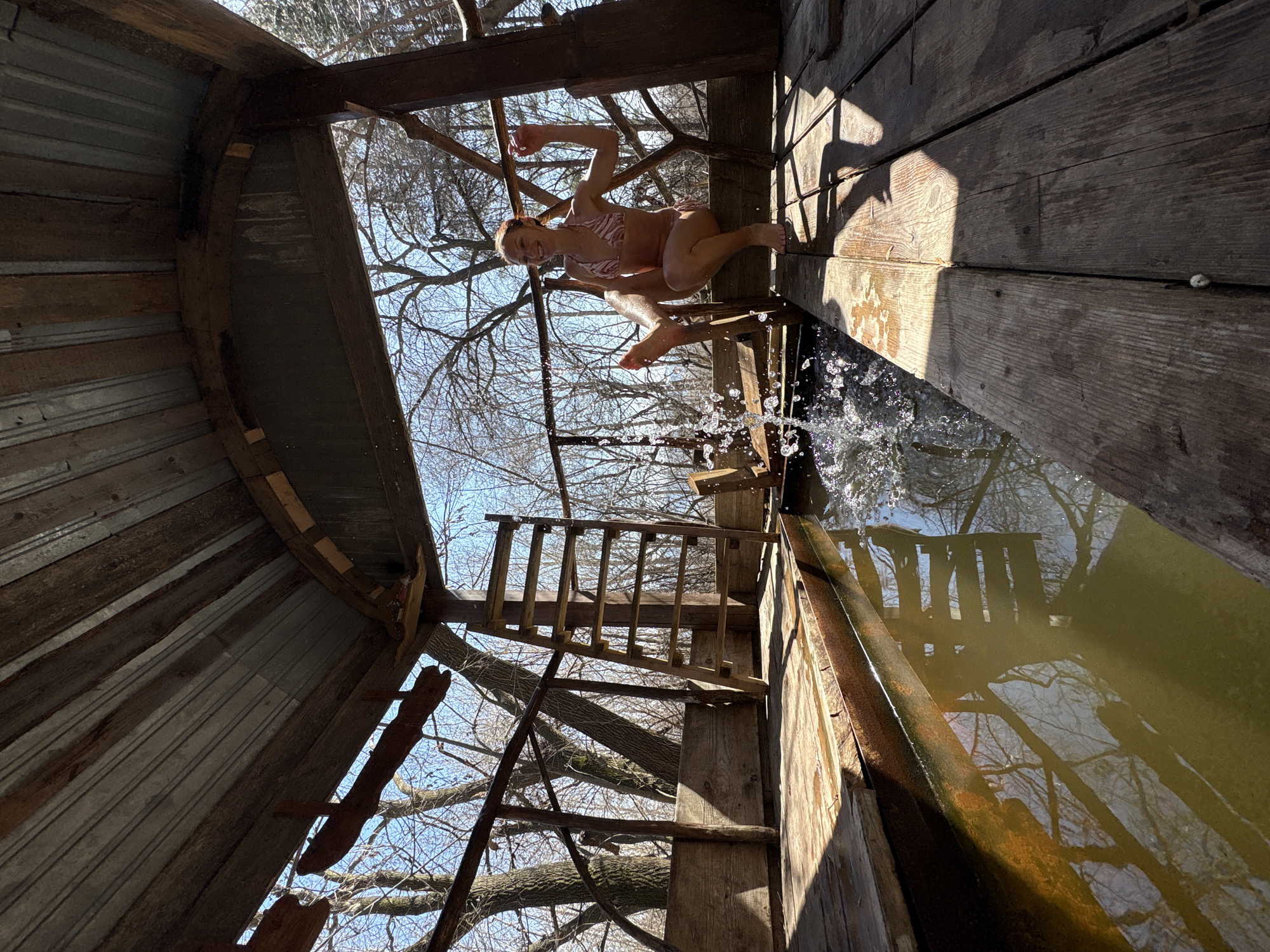 A woman is leaping from a rustic wooden platform into a water-filled trough. The platform, constructed of aged lumber, is part of a simple structure with walls made of corrugated metal and wood, partially open to the sky. The woman is fair-skinned and wearing a pink and white patterned bikini. Her body is arched as she is suspended in mid-air, with water splashing from the edge of the platform as she jumps. A crude wooden ladder leads up to the platform, and branches of trees are visible through the open roof. The water in the trough reflects the sky and surrounding trees. The location is not discernible from the image, and there is no evidence to support that it is located in Ulyanovsk, Russia.