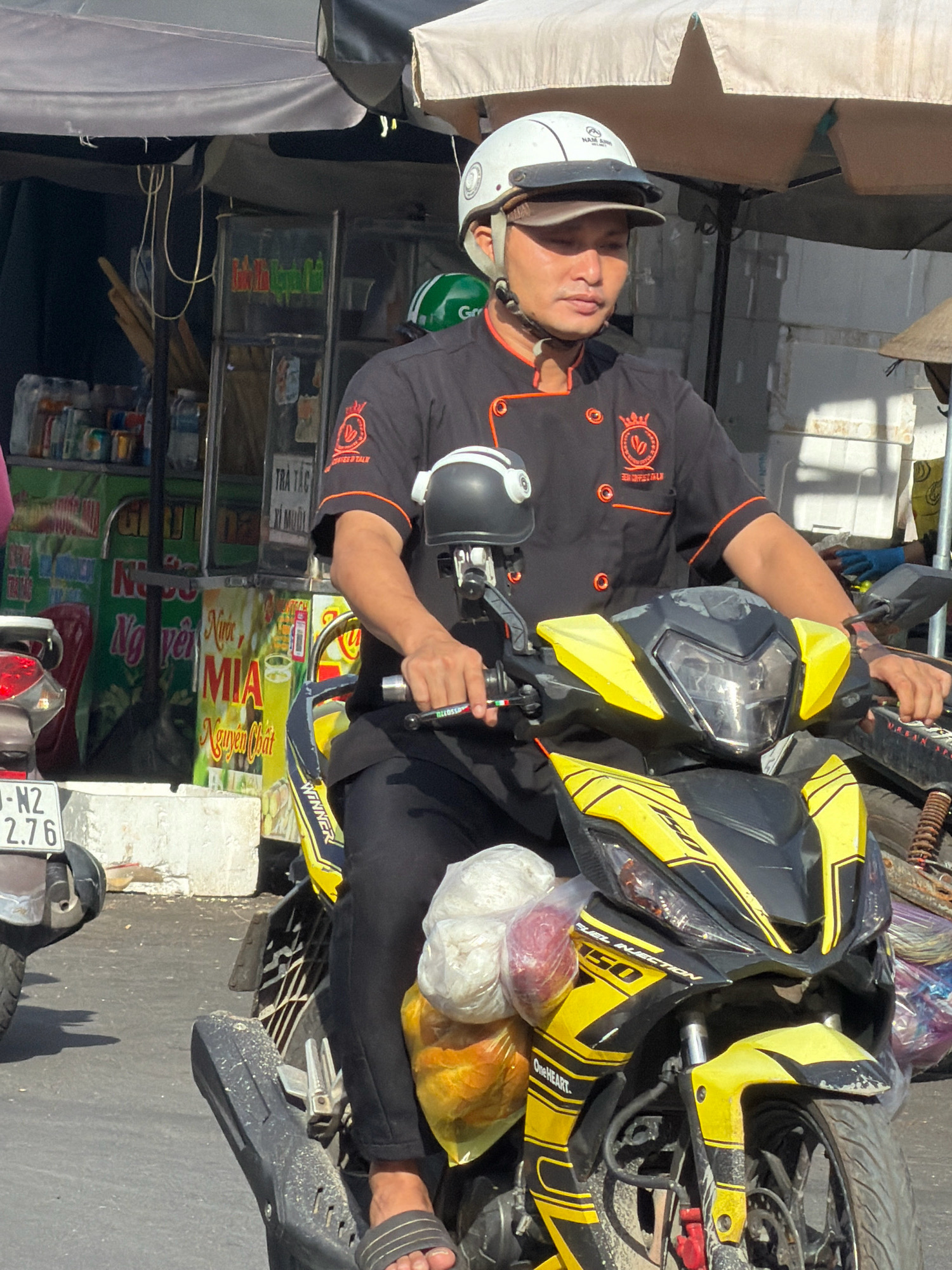 A man wearing a black chef's uniform with orange trim and a white helmet is riding a yellow and black motorcycle. He is holding the handlebars with his right hand and the clutch lever with his left hand. The motorcycle is parked on a street with a market stall in the background. The market stall has various items for sale, including drinks and food. The man appears to be a delivery driver or vendor. The location is a street in Nha Trang, Vietnam, evident from the Vietnamese writing on the market stall signage.