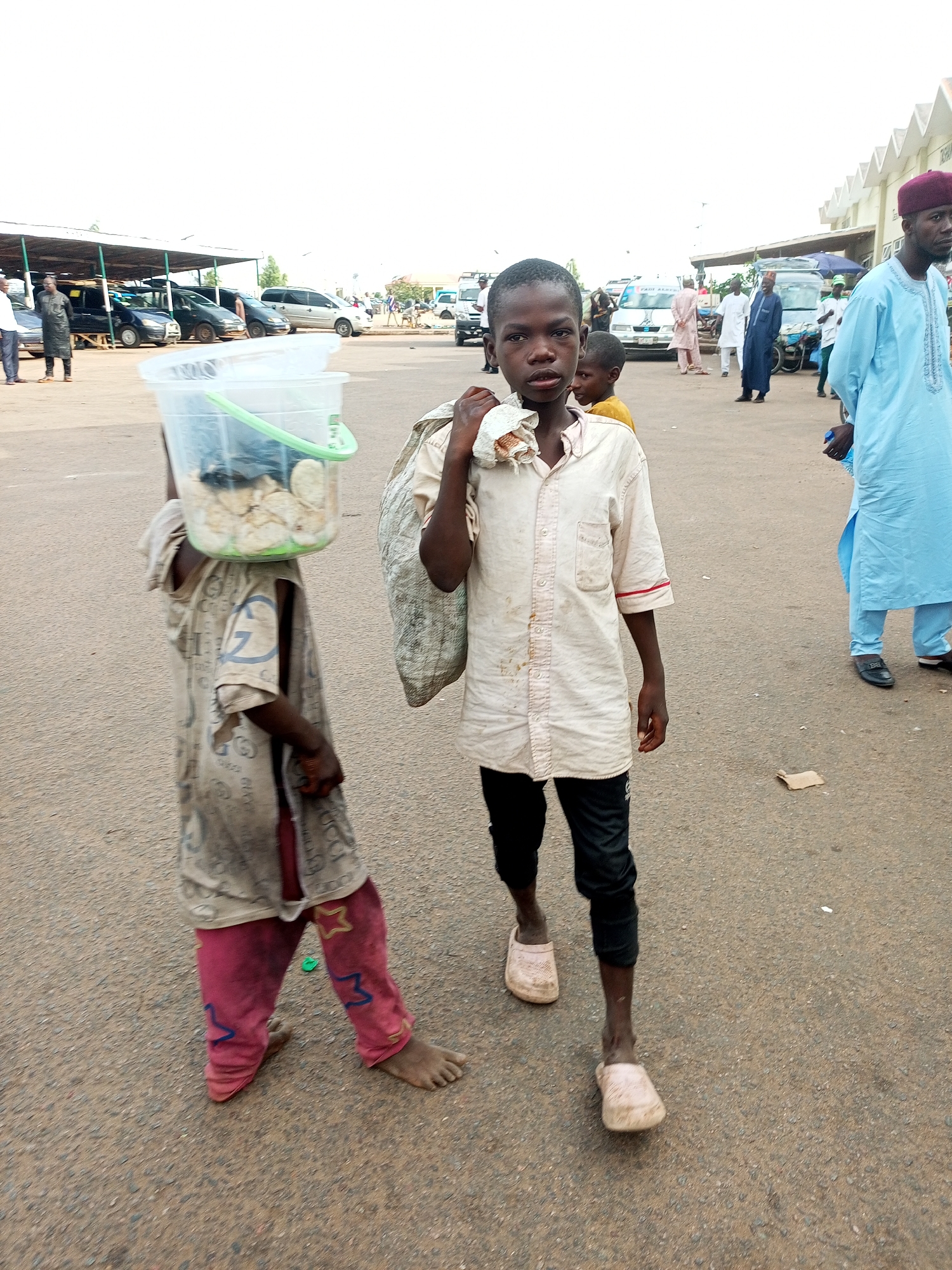 In the city of Gombe, Nigeria, two young boys are walking on a paved surface. The boy in the foreground, on the right, is looking at the camera and wearing a cream-colored shirt with red trim on the sleeves, and black pants. He is holding a large, woven bag over his right shoulder. The boy on the left is walking away from the camera and is carrying a clear plastic bucket filled with what appears to be food items on his head. He is wearing a tan, patterned t-shirt and pink pants decorated with stars and abstract shapes. In the background, several vehicles are parked under a covered area, and other people are visible walking around.