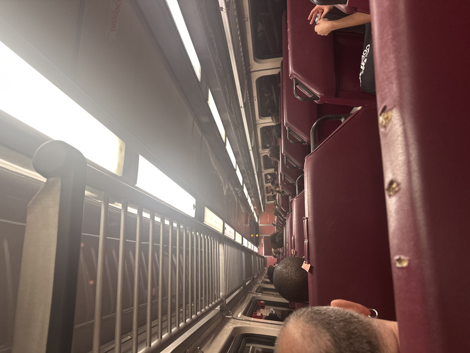 The image captures the interior of a passenger train car, identified as operating within or passing through Wellesley, United States. The perspective is from a seated passenger, looking forward down the aisle.
The foreground prominently features the back of a male passenger's head, partially bald with gray hair, wearing a silver hoop earring in the left ear. The maroon upholstered seat directly in front of the camera exhibits minor surface wear. Beyond this, rows of identical maroon seats extend down the train car. Several other passengers are visible, primarily from behind, occupying various seats. On the right side, the hands of a passenger are visible, holding a small, dark object.
The ceiling of the train car is white, equipped with multiple elongated fluorescent light fixtures that provide bright, overhead illumination. A metallic, grid-patterned luggage rack runs horizontally along the upper left side of the car. An "EMERGENCY" sign, rendered in red text, is visible on a ceiling panel towards the upper right. Windows line both sides of the car, appearing dark, which obscures any exterior view. The environment is orderly, indicating active transit with multiple individuals present in a seated, non-interacting state.