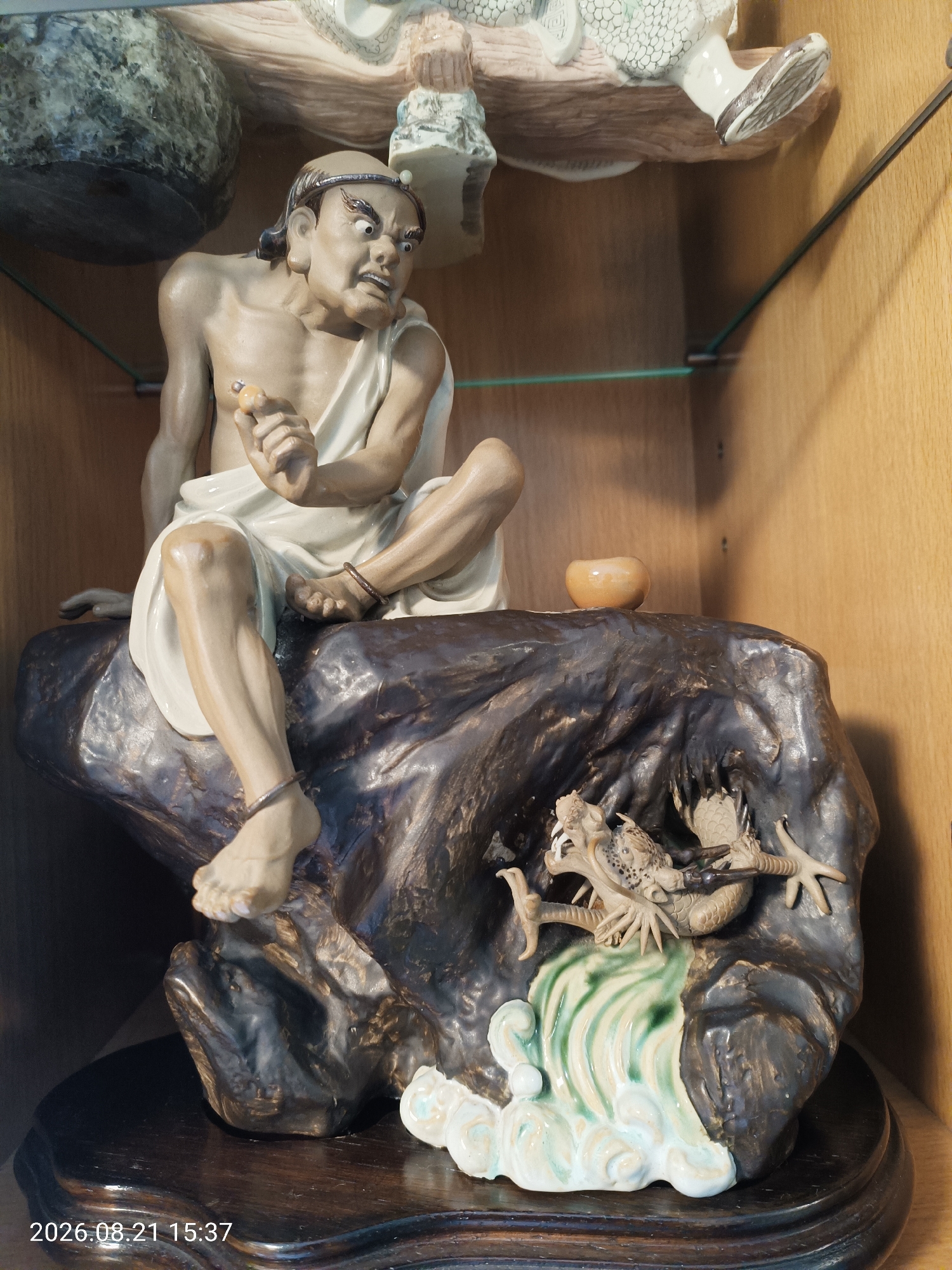 A detailed figurine depicts a person sitting on a dark, rocky surface. The figure has a distressed expression and is clad in a white cloth draped over their body. They are holding a small, light-colored object in their right hand. The rocky surface features a dragon coiled at its base, with green and white waves cascading around it. A small, round, orange bowl rests on the rock behind the seated figure. The figurine is displayed on a dark, polished wooden base within a wooden cabinet. The timestamp "2026.08.21 15:37" is visible in the lower left corner of the image. The setting appears to be indoors, possibly a home or a display cabinet in a shop. There is no specific indication of a location in Hong Kong, China, as the scene is a static display.