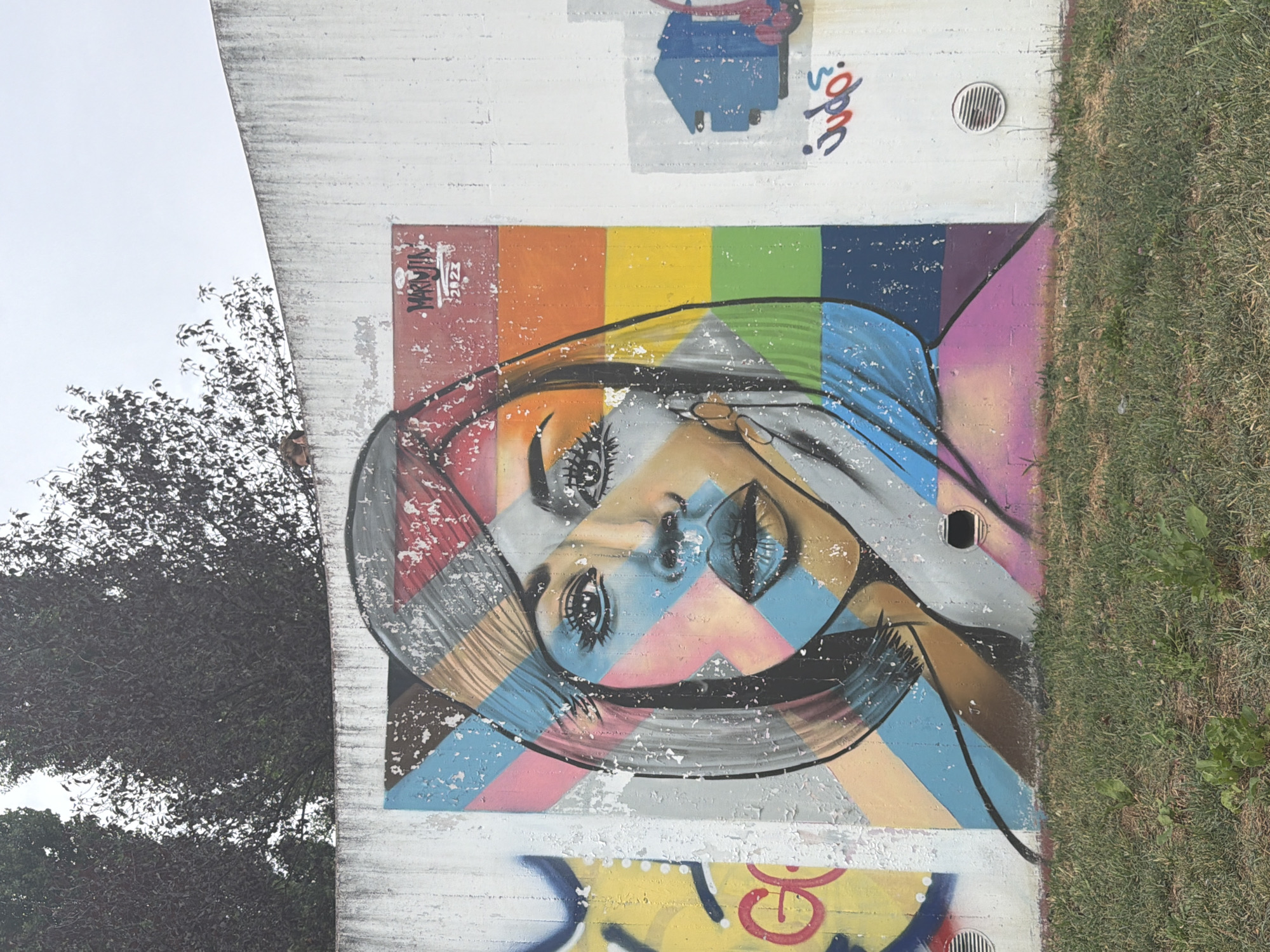 The image depicts a large, colorful mural painted on a rough, weathered concrete wall. The mural features a stylized portrait of a woman with striking eyes and blue lips. Her face is overlaid with geometric shapes in various colors, including pink, blue, yellow, and red, creating a mosaic-like effect. To the right of the portrait, a section of the wall displays a rainbow-colored stripe pattern. Above the mural, near the top right corner, the artist's tag "MARMJIN" and the year "2023" are visible. To the left of the main mural, parts of another graffiti piece are visible, including yellow paint and red lettering that appears to read "GO". To the right of the main mural, there is a smaller graphic of a blue house-like structure and some stylized text that appears to read "tudo?". The wall is situated outdoors, with a grassy area in the foreground. Tall, dark trees are visible behind the wall against a cloudy sky. The overall scene appears to be in an urban environment. This mural is located on the wall of the former market in Corsico, Italy.