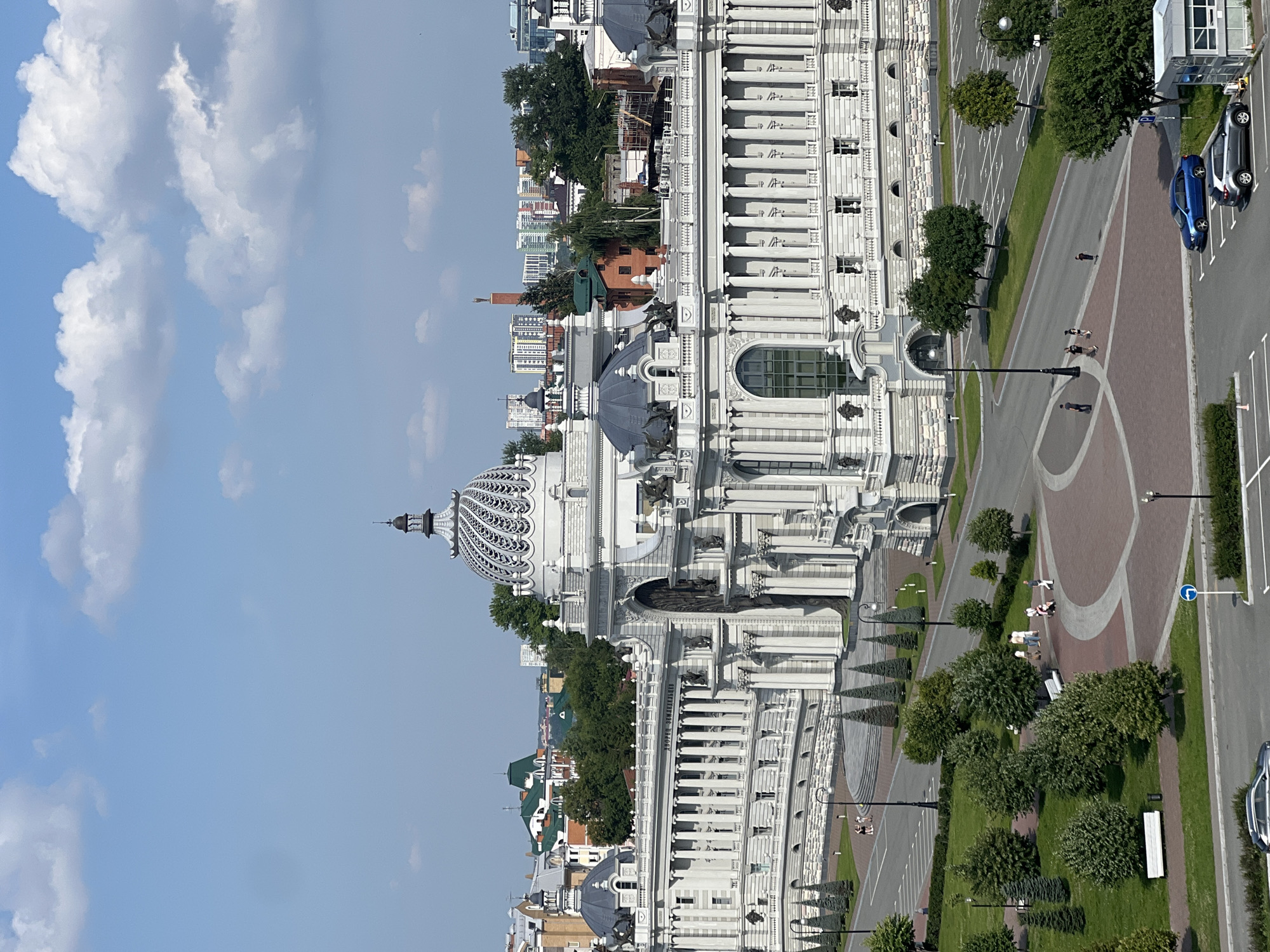 The image shows the Palace of Agriculture in Kazan, Russia, on a clear day with some clouds in the sky. The grand white building features ornate columns, multiple domes, and intricate architectural details. In the foreground, there is a green lawn with trees and walkways, where several people are strolling. A few cars are parked in designated spots on the pavement. The background reveals more of the city, including residential buildings and a distant chimney.