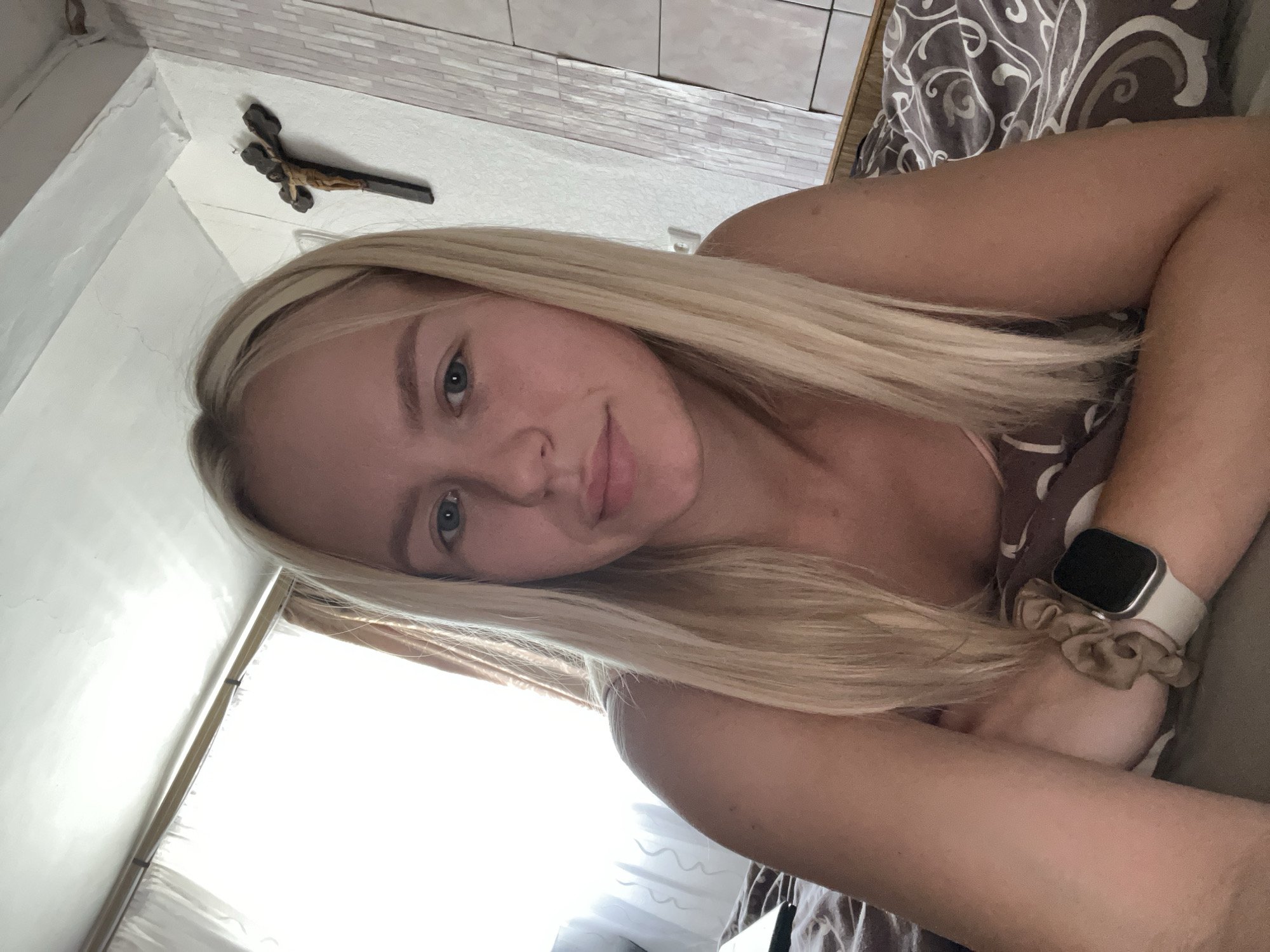 The media file shows a young Caucasian woman with long blonde hair, lying down and looking at the camera. She is wearing a light-colored shirt and a watch on her left wrist. Her hair covers part of her face and body. A crucifix is visible on the wall behind her, and a patterned blanket or comforter is in the background. The lighting suggests it might be daytime, with natural light coming from a window to the left of the frame. The woman's expression appears relaxed and perhaps a little sleepy. The overall scene is intimate and personal, likely taken in a bedroom or similar private space. There is no visible text in the image.