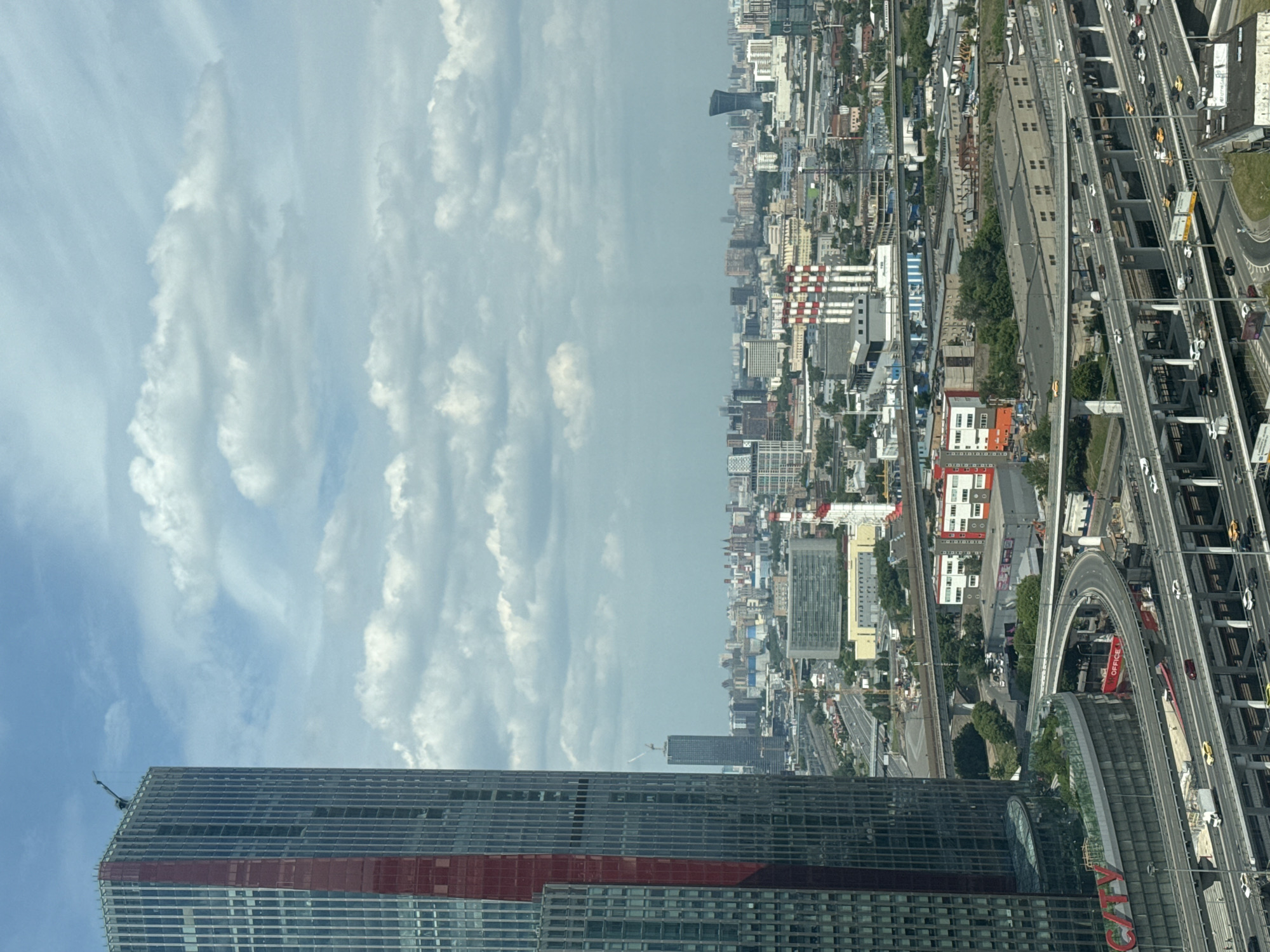 This is an eye-level, medium shot of a vast, sprawling urban landscape under a partly cloudy sky. The foreground is dominated by the imposing, glass-clad skyscraper with a striking red vertical stripe, on the left side of the frame. A construction crane is visible at the apex of this building. Below the skyscraper, a complex, multi-layered highway interchange bustles with traffic, including cars, trucks, and a red truck. The highway infrastructure features numerous concrete pillars and elevated road segments.
Beyond the immediate vicinity of the skyscraper and highways, the cityscape extends into the distance, revealing a dense collection of diverse buildings. Numerous industrial structures, including rows of large warehouses and buildings with visible pipes and chimneys, are interspersed with office buildings and residential blocks. Prominent features in the mid-ground include a cluster of red and white-striped chimneys, likely part of a power plant or industrial facility. Further in the background, a distinctively shaped, dark skyscraper with a curved facade stands out against the horizon. The urban sprawl appears to extend as far as the eye can see, suggesting a large metropolitan area. The overall impression is one of a densely populated and industrialized city.
The image depicts the city of Khimki, Russia, specifically showcasing the Moscow City business district and its surrounding areas, including infrastructure like the MKAD (Moscow Ring Road) and industrial zones.