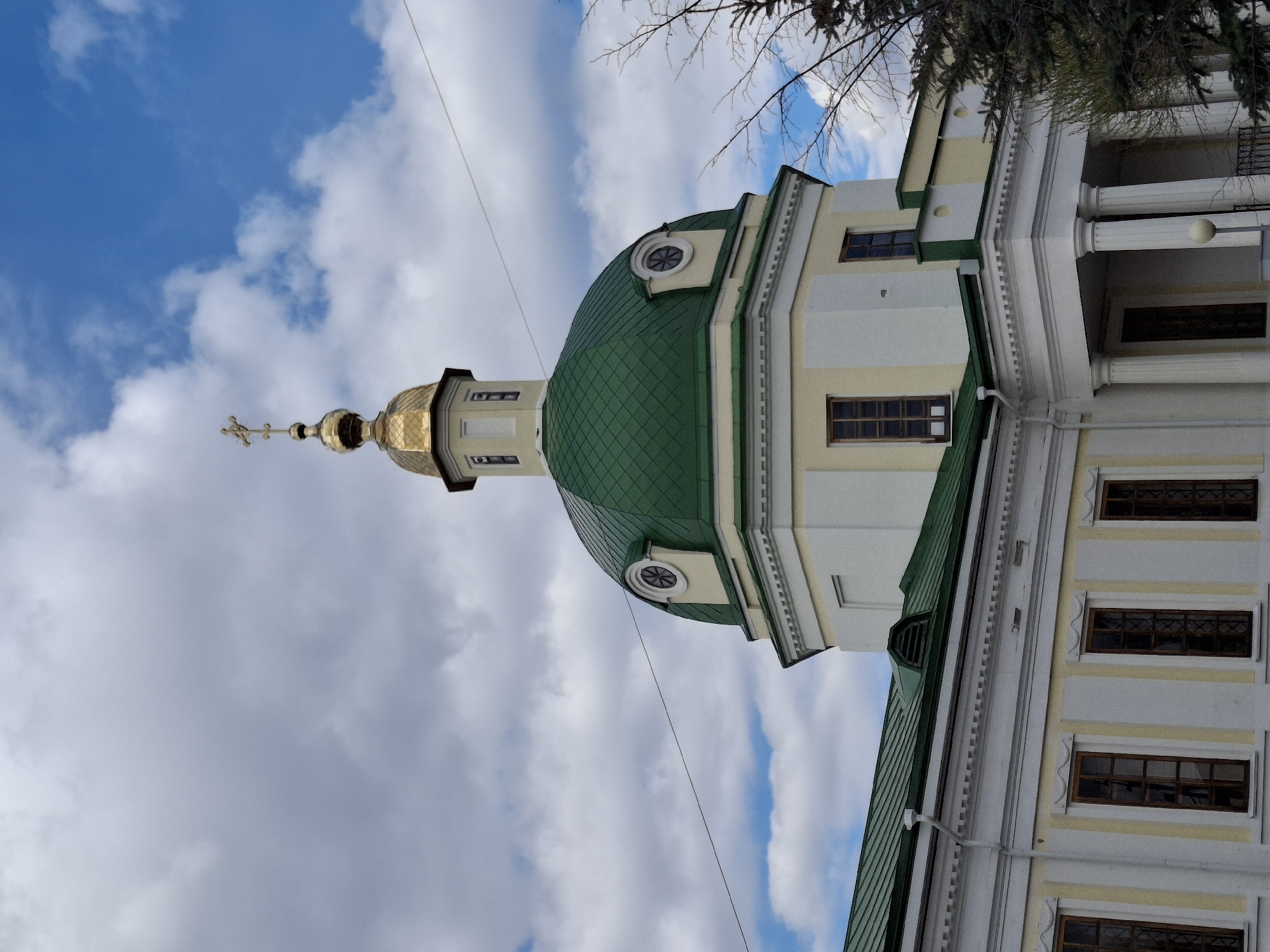 An upward-angled outdoor shot captures the intricate architecture of a religious building, likely an Eastern Orthodox church, under a bright, partially cloudy sky in Sarapul, Russia. The most prominent feature is a large, green, tiled dome, from which a narrower, octagonal tower rises, topped by a smaller, golden onion-shaped dome. This golden dome is crowned with a distinctive golden Orthodox cross.
The building's walls are painted in alternating sections of light cream and white, with architectural detailing such as pilasters and cornices. Several dark-framed windows are visible along the building's facade, and a circular window adorns the base of the green dome. On the right side, a section of the building features classical columns, suggesting a grand entrance or portico.
In the upper right corner, bare tree branches are visible against the sky, indicating a colder season such as late autumn or winter. A thin, dark wire traverses the blue sky diagonally from left to right, passing in front of the building. The scene is brightly lit by natural daylight, with patches of blue sky and scattered white clouds. No people or specific activities are depicted, focusing solely on the architectural subject. No visible text is present.