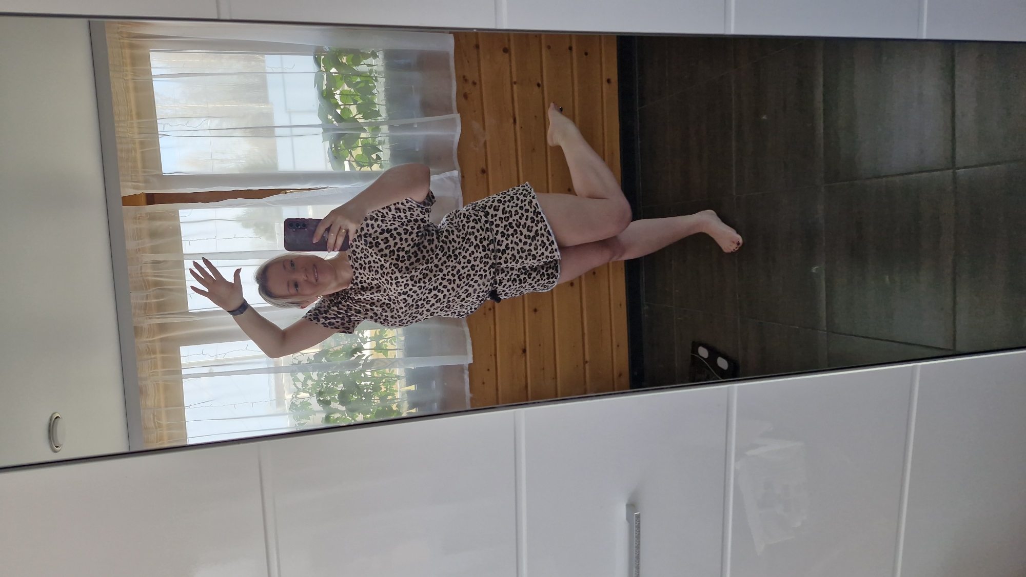 A woman is reflected in a mirror, taking a selfie. She has short blonde hair and wears a leopard-print dress. She is holding a phone in her right hand and waving with her left. The reflection shows a room with wood paneling, a window with sheer curtains and plants, and dark-colored tiles on the floor. The mirror is framed by white tiles or panels. The setting is likely a domestic interior, possibly a bathroom or bedroom. The location is Otchor, Russia.