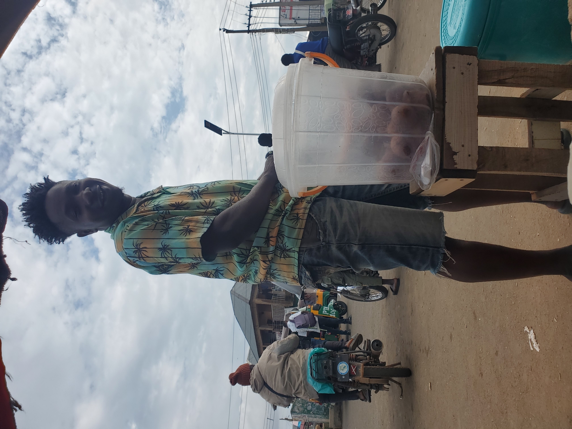 A man with dark skin and short, textured dark hair smiles at the camera while standing on a dirt road in Gwong, Nigeria. He wears a short-sleeved, collared shirt with a palm tree pattern in green, yellow, blue, and orange. His lower body is out of frame, but he appears to be wearing denim shorts. To his right, a clear plastic bucket filled with round, brown objects sits on a wooden stool. The bucket has orange handles and a white lid. In the background, a woman with a brown knitted hat rides on the back of a motorcycle, facing away from the camera. Other motorcycles and a three-wheeled vehicle are also visible, along with buildings and utility poles. The sky is cloudy.