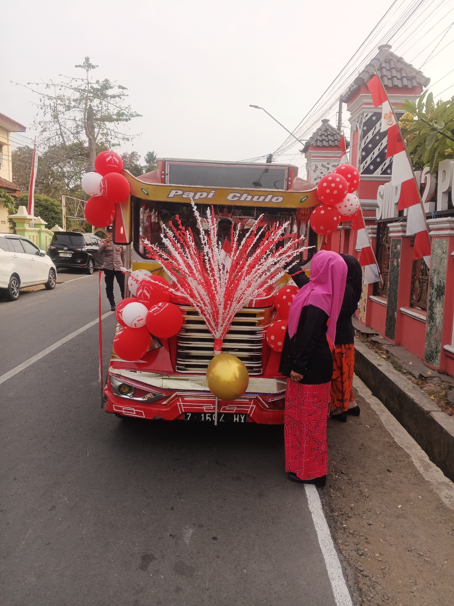 A red and yellow decorated bus, likely part of a parade or celebration, is parked on a paved road in Sokanegara, Indonesia. The bus is adorned with numerous red and white balloons, some with polka dots, and a prominent red and white decorative spray. A golden balloon is positioned on the front of the bus. The bus's license plate reads "Z 1507 HX". Two individuals are visible near the rear of the bus: one in a pink hijab and patterned skirt, and another person partially obscured. A police officer in uniform is standing in the road to the left of the bus, holding a mobile phone. In the background, a building with red and white decorative elements, including Indonesian flags, is visible. A white car and a black SUV are parked on the side of the road. The sky is overcast.