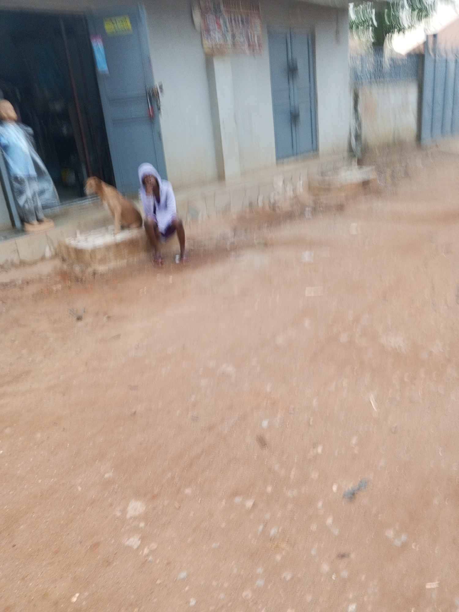 Outside a simple concrete building with blue doors, a person in a white hooded sweatshirt sits on the steps beside a brown dog. The surrounding ground is reddish-brown dirt. The location is in Abuja, Nigeria.