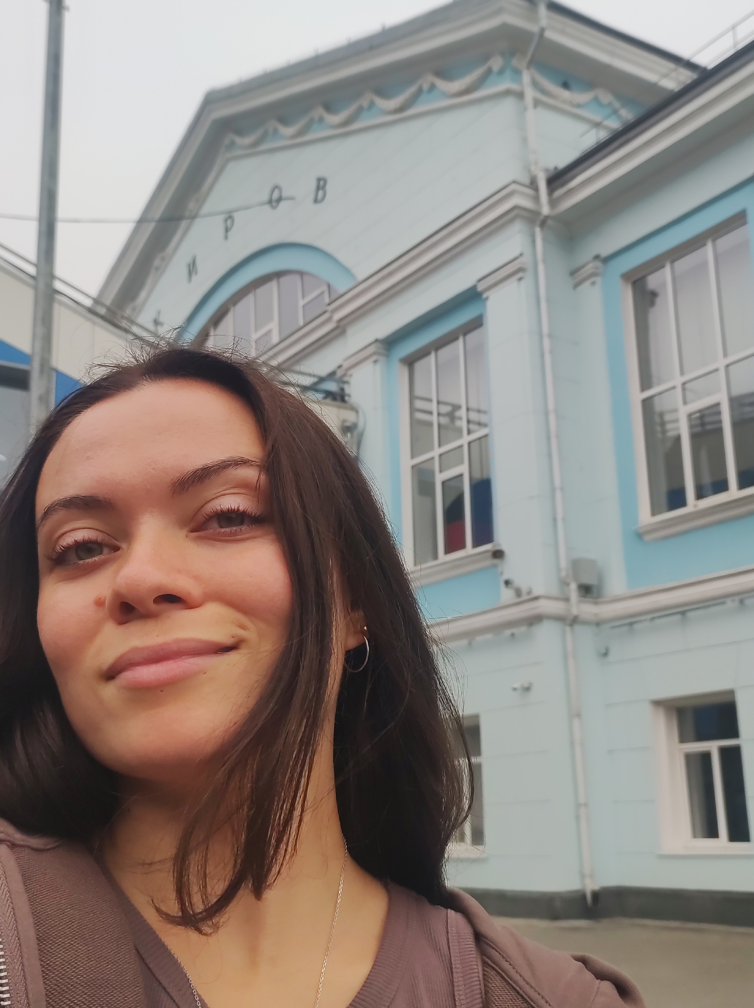 A woman is taking a selfie outside a light blue building with white trim and decorative elements. The building has several windows with white frames and internal grids. Above the windows, there are inscriptions in Russian that appear to read "КИРОВ" (KIROV). The sky is overcast. The woman has dark brown hair and is wearing a brown zip-up hoodie and a silver necklace. She is smiling slightly. The image appears to be taken outdoors in the city of Kirov, Russia, in front of a building that likely serves a public function.