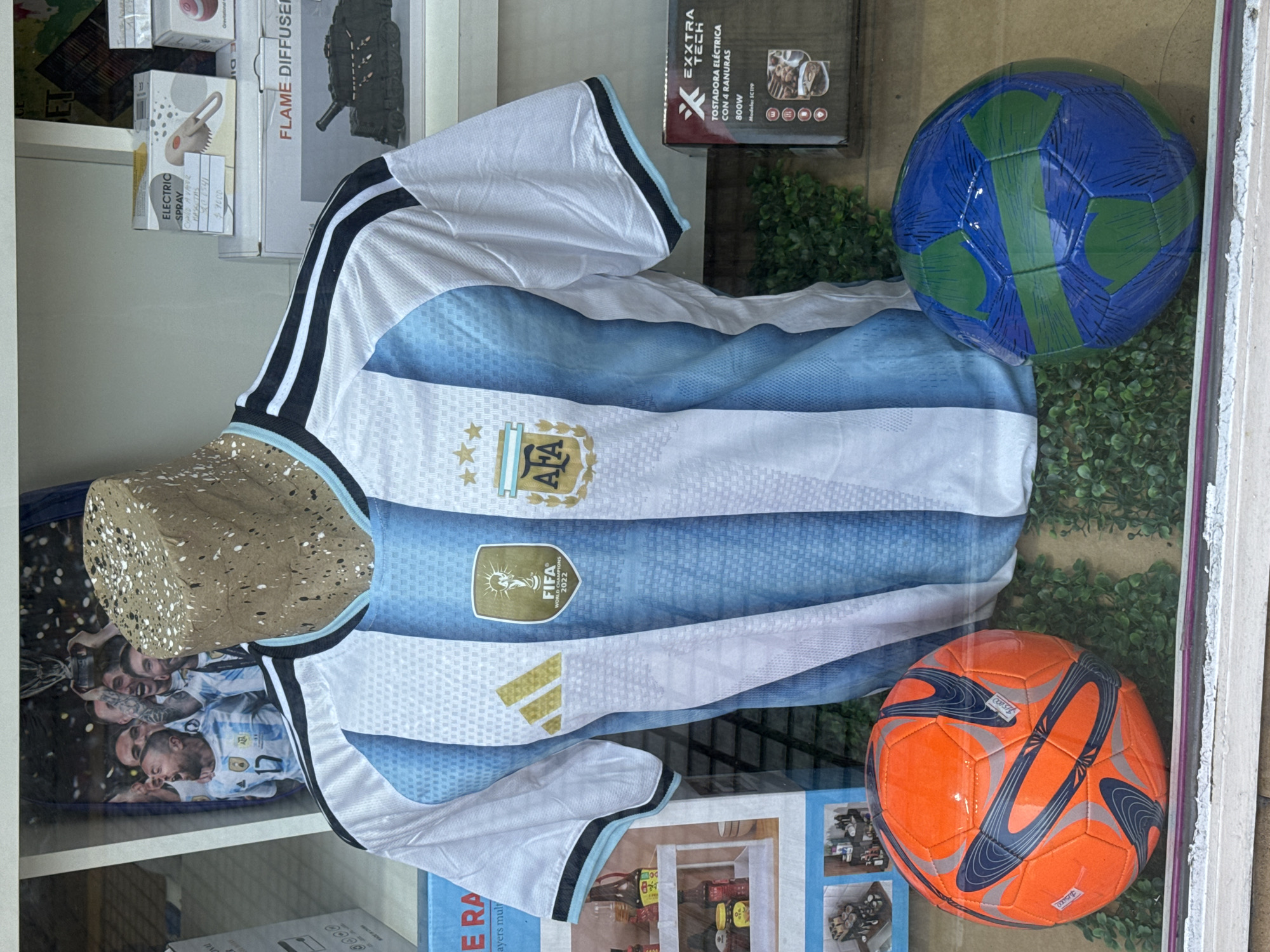 The image shows a storefront display in what appears to be Buenos Aires, Argentina. The focal point is a white and light blue striped Argentina national football team jersey, with the AFA (Argentine Football Association) crest and FIFA World Champions 2022 badge prominently displayed. The jersey is on a mannequin of some kind. To the right of the jersey are two soccer balls, one with a blue and green design, and the other orange with gray accents.
In the background and along the sides, various retail items are visible on shelves, including a box labeled "FLAME DIFFUSER," a box labeled "EXXTRA TECH TOSTADORA ELECTRICA," and a box labeled "ELECTRIC SPRAY CUPID AVATOR MASCOTAS." A poster featuring the Argentinian football team is also partially visible. The price of the spray is 7,000. A sticker or tag is on the orange ball saying $15000.