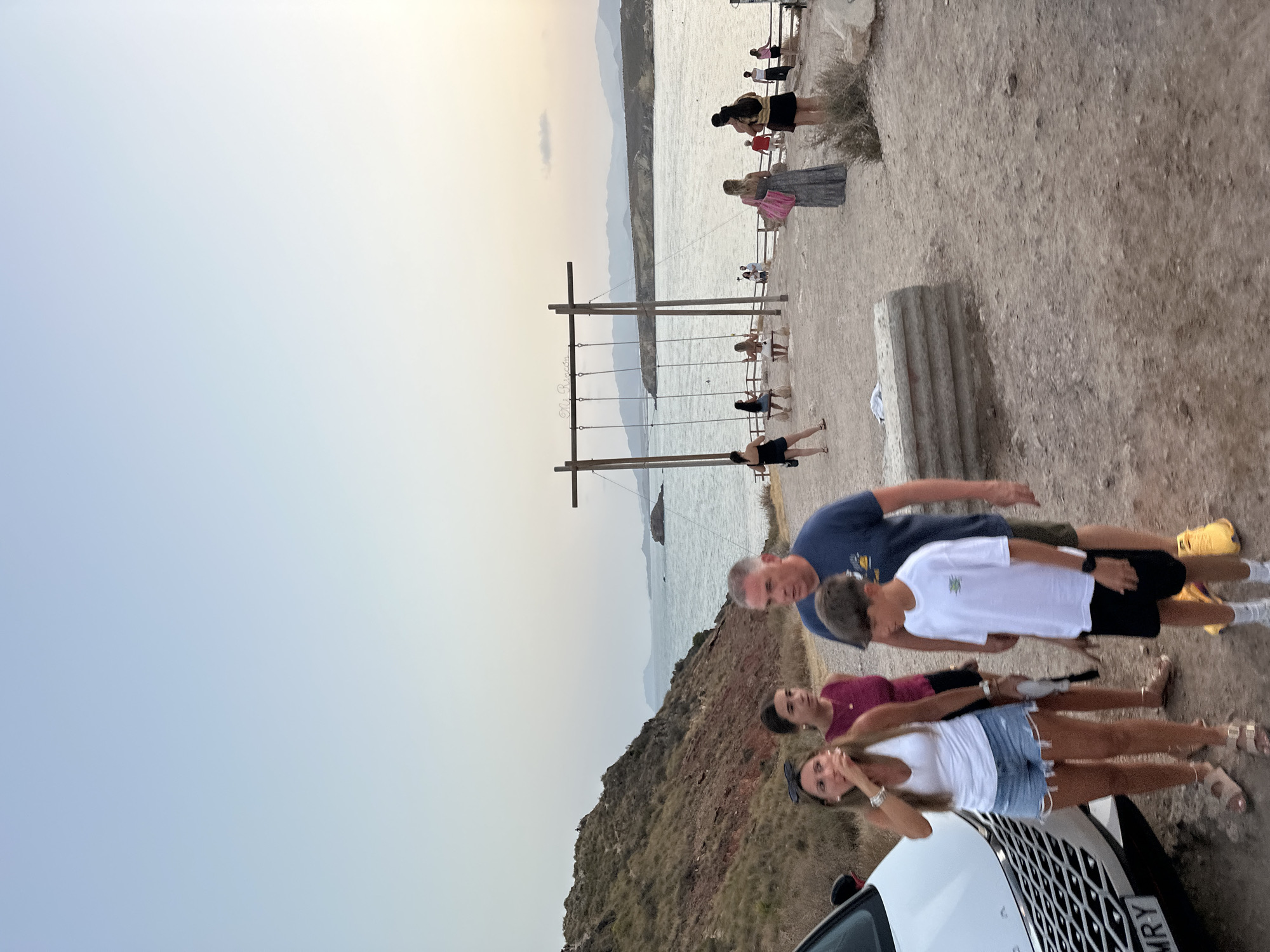 The image depicts a coastal scene at dusk in Mazarrón, Spain. In the foreground, a family of four, consisting of two adults and two children, are walking on a dirt path. The woman in front is wearing a white tank top and denim shorts, and the man beside her is in a navy blue t-shirt. Behind them, a younger woman in a maroon top and a boy in a white t-shirt and black shorts are also visible. To the left, a white car is partially visible.
In the background, a scenic view of the Mediterranean Sea unfolds, with hills and mountains forming the distant coastline. A prominent feature is a large wooden swing set structure with three swings suspended over the sandy ground. Several other people are scattered around the area, some are near the swings, while others are further back along a wooden fence. The lighting suggests it is late afternoon or early evening, with a soft, hazy sky. The overall atmosphere is relaxed and recreational, indicating a popular spot for visitors.