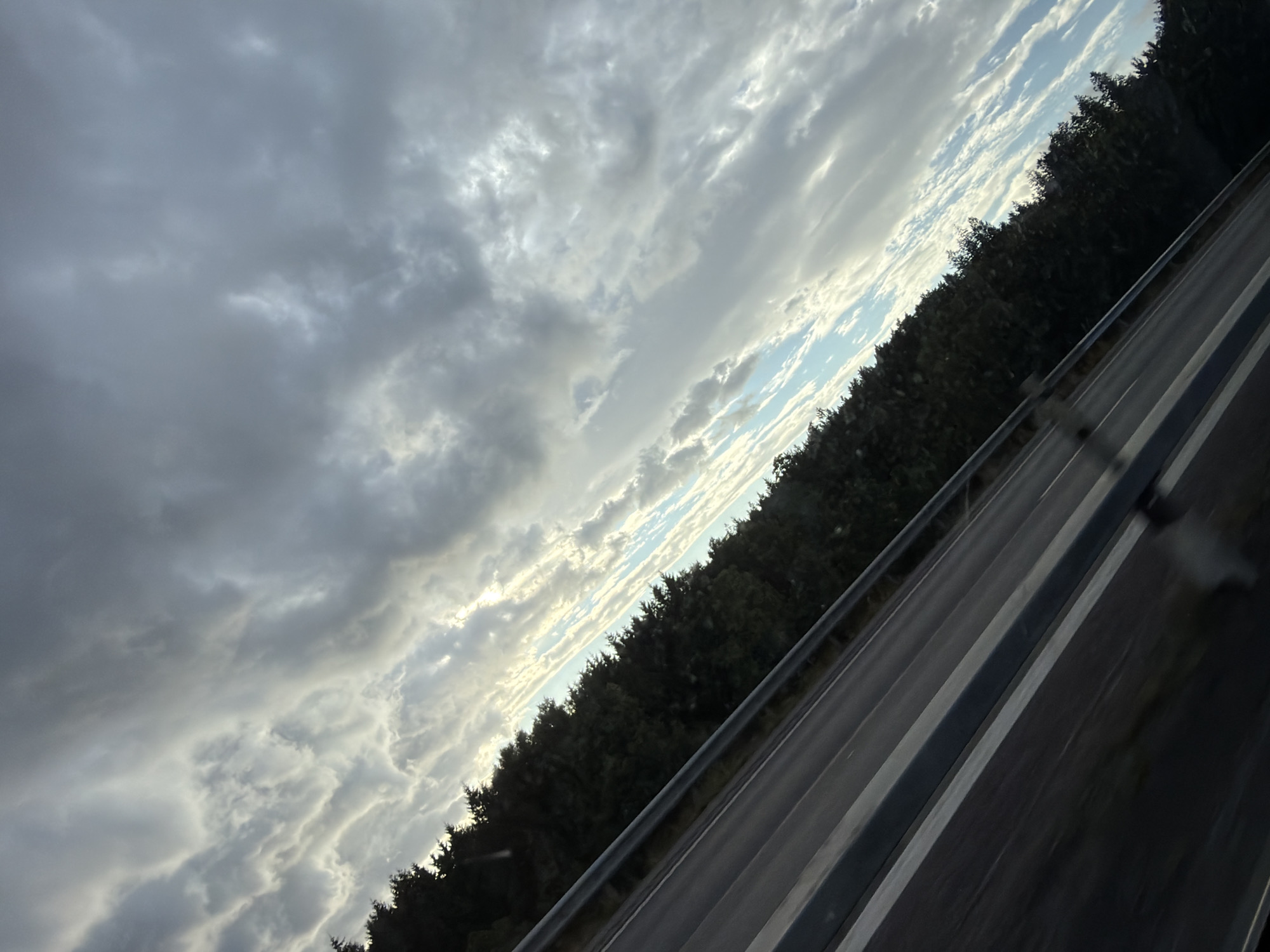 This image captures a view from a moving vehicle on a highway, likely in or near Löftaskog, Sweden. The perspective is angled, looking towards the sky and a dense line of dark green trees bordering the road. The sky is dominated by dramatic, gray and white clouds, with patches of blue sky visible between them. Sunlight is breaking through in some areas, creating highlights on the clouds. The highway itself is visible in the foreground, with dark asphalt and white lane markings. A metal guardrail runs along the side of the road. The overall impression is one of a somewhat overcast and dynamic sky, seen from a typical roadside environment.