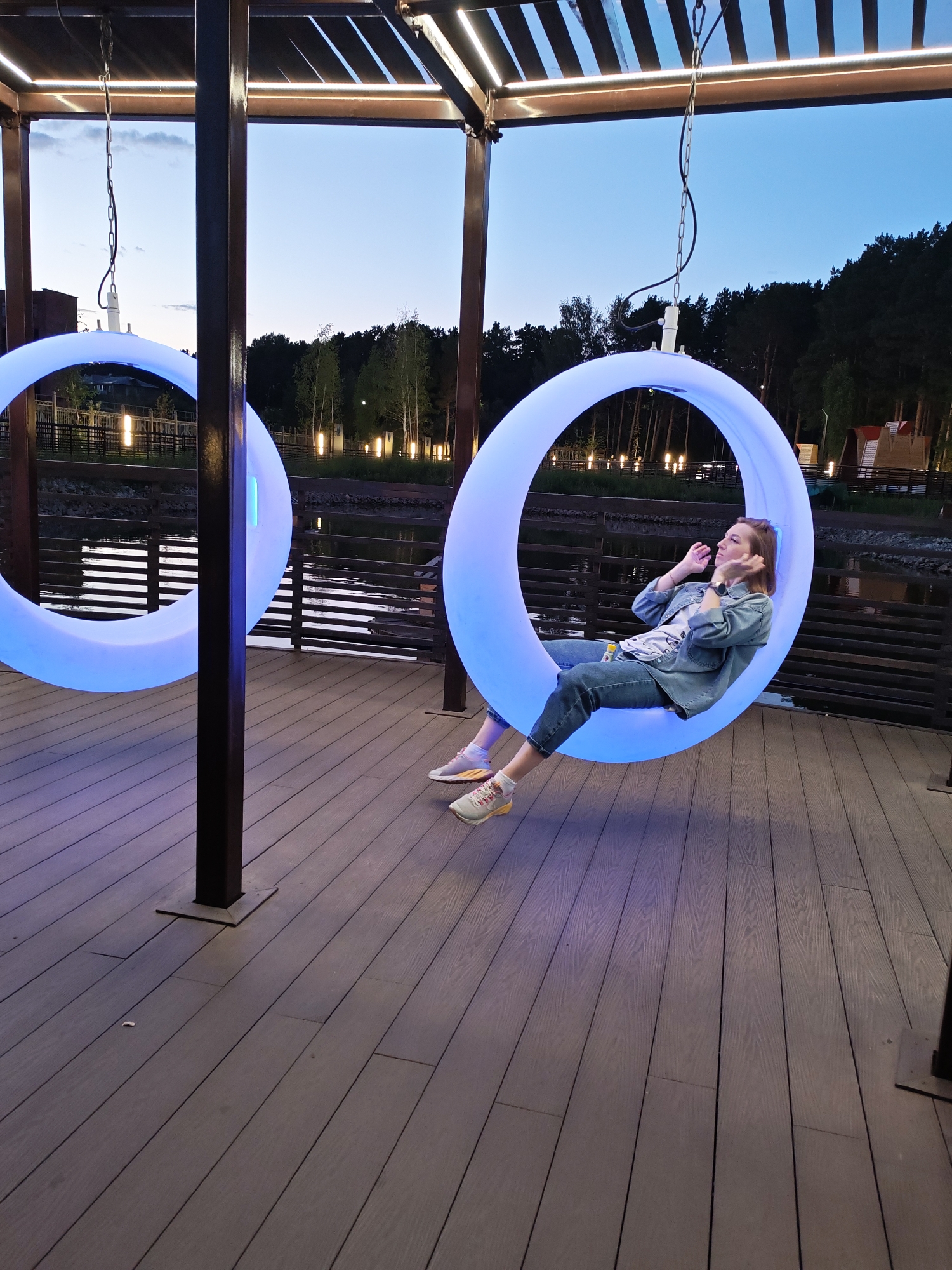 A woman is sitting on a large, circular, illuminated swing, which is hanging from a pergola. The swing is lit with a cool blue light. She is wearing a denim jacket, jeans, and sneakers, and her hair is pulled back. Her arms are resting on her lap, and her gaze is directed to her right. The pergola has a slatted roof and wooden beams. The floor beneath the pergola is made of wooden planks. In the background, there is a body of water, a wooden fence, trees, and some lights. The sky is a deep twilight blue, suggesting it is evening. The scene is outdoors, likely a recreational area. This location is identified as being in Seversk, Russia.