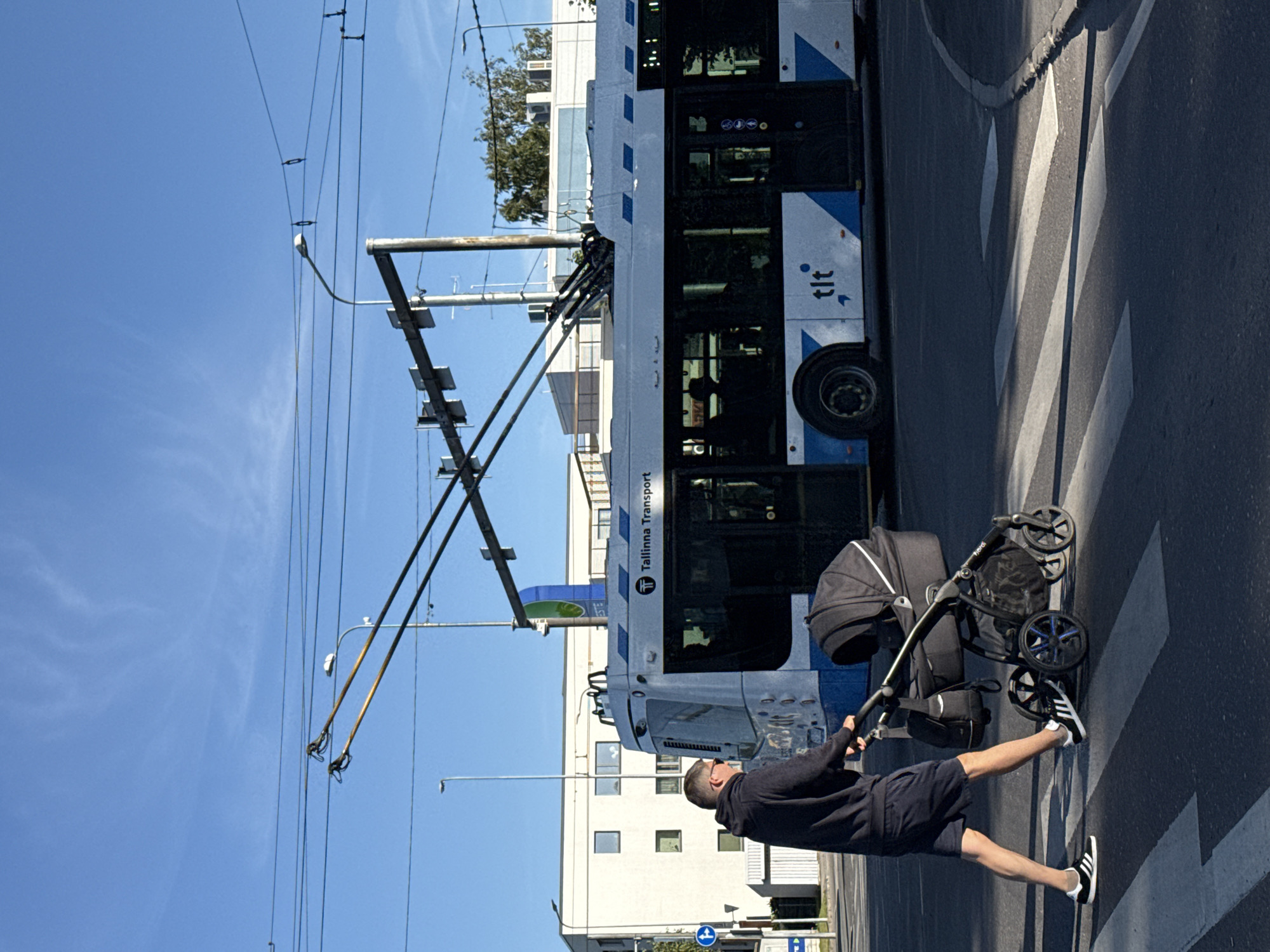 A man, wearing a dark hoodie and navy blue shorts, pushes a black stroller across a pedestrian crossing. He is wearing black and white athletic shoes. The stroller contains a dark gray bassinet. A white and blue trolleybus with "Tallinna Transport" written on its side is stopped next to the pedestrian crossing. In the background, there is a modern white building and a clear blue sky with some overhead wires and streetlights. The image appears to be taken in Tallinn, Estonia, given the presence of the "Tallinna Transport" branding on the trolleybus.