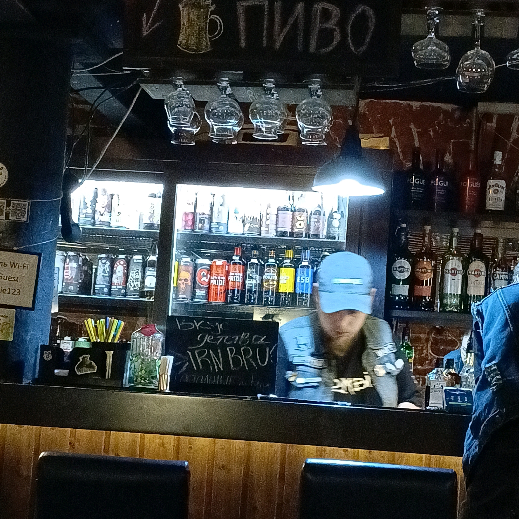 The image depicts the interior of a bar, likely in Samara, Russia, given the Cyrillic text visible. A bartender, wearing a blue baseball cap and a denim jacket, is partially visible behind a counter. The counter has a chalkboard with Cyrillic writing that appears to be a menu item, "Вкус... делава IRN BRU!". Several bottles of alcohol are displayed on shelves behind the counter, including recognizable brands like Martini and Jim Beam. Various glassware, such as wine glasses, are hung upside down above the shelves. The bar area appears dimly lit with some ambient lighting from above and the illuminated shelves. On the left side of the counter, there is a sign with text, including "Wi-Fi" and "Guest", and a QR code. The overall atmosphere suggests a casual, perhaps slightly rustic, bar setting.
