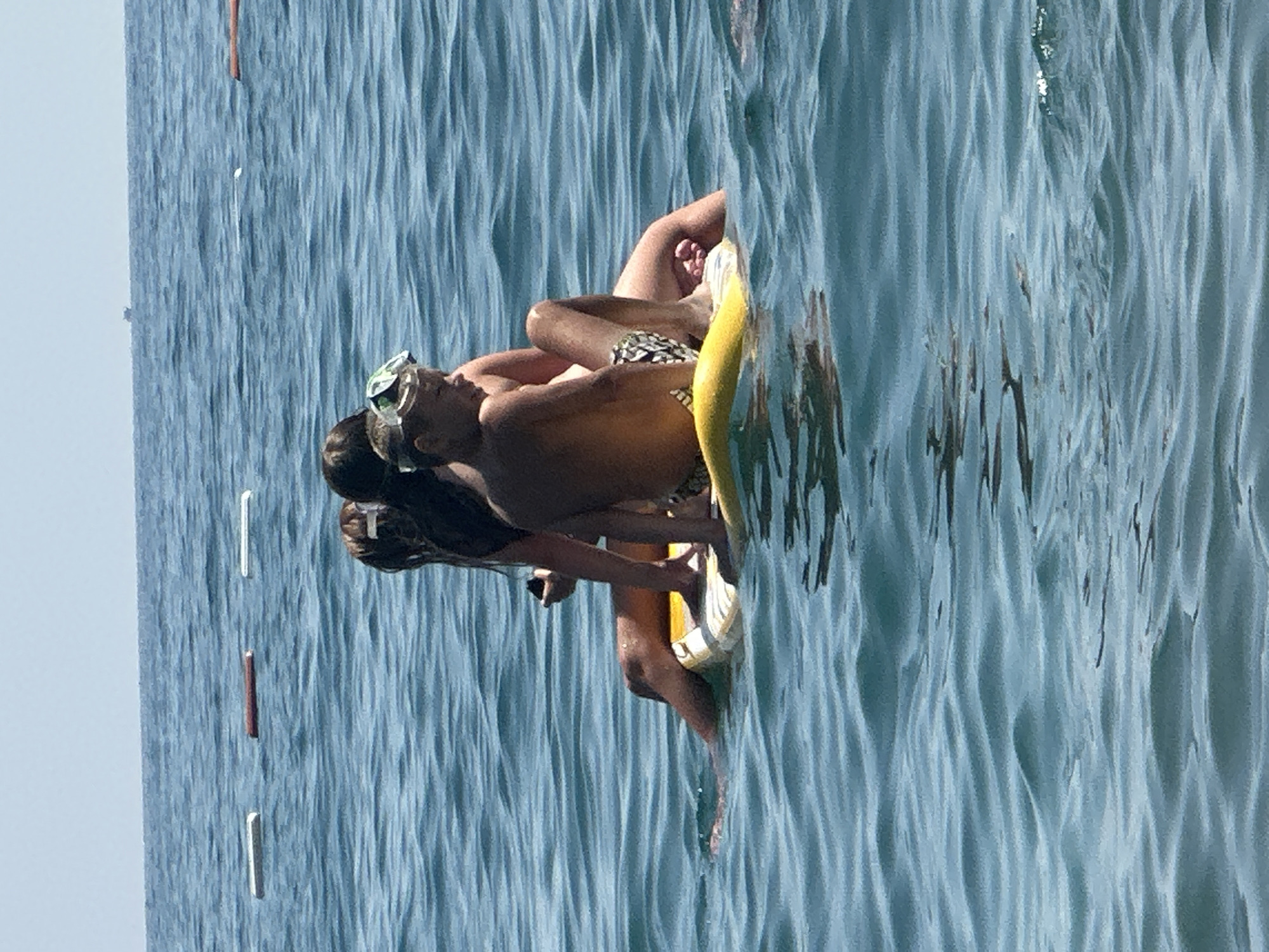 In the city of Boreti, Montenegro, four individuals, including two young boys and two young girls, are seated on a yellow flotation device in the blue, wavy sea. The children appear to be enjoying a sunny day outdoors. The background shows a clear sky and the vast expanse of the sea, with a few buoys visible in the distance. The overall scene depicts a recreational moment on the water.