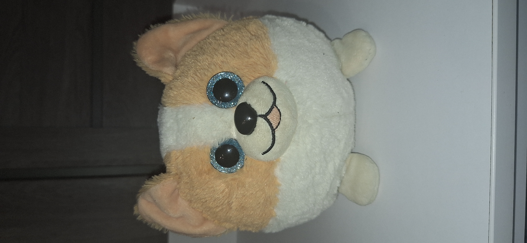 A plush toy dog, likely a corgi, sits on a white surface. The toy is round with fluffy tan and white fur. It has large, sparkling blue eyes and a black nose. Its ears are tan and stand upright. The background is dark and appears to be a wall or furniture. The scene is indoors.