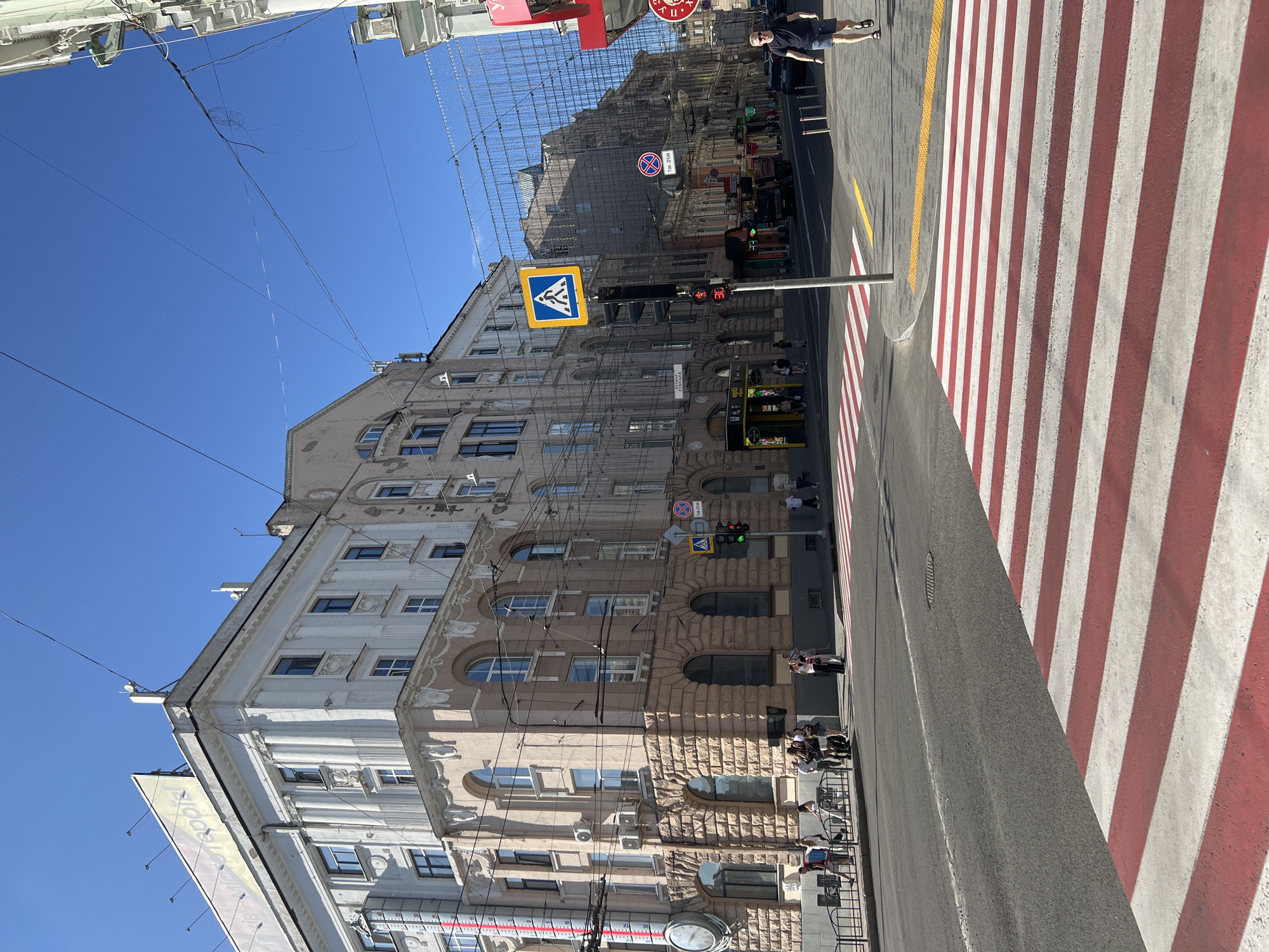 The image captures a sunny day on a street corner in Kharkiv, Ukraine, focusing on a large, ornate building with a light pink facade and a white upper story. A prominent red and white striped crosswalk dominates the foreground, indicating a pedestrian crossing. Across the street, a man in a dark t-shirt and shorts walks away from the camera.
The building features classical architectural elements, including arched windows, columns, and decorative moldings. A large analog clock is visible on the side of the building, near a tall red and white striped structure that appears to be a thermometer. Several people are gathered on the sidewalk near the building, some walking and others standing in groups.
A pedestrian crossing signal with a yellow diamond sign depicting a walking figure is mounted on a traffic light pole. The traffic light is illuminated red. Further down the street, other buildings line the road, and power lines and decorative string lights are suspended overhead. Signs with Cyrillic script are visible on the buildings and poles. The overall scene suggests a busy urban environment on a clear day.