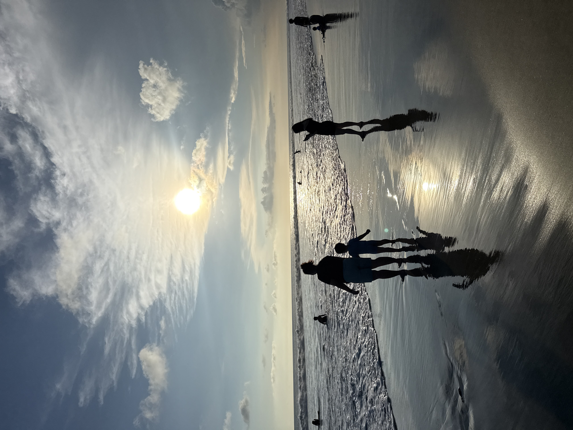 This media file captures a serene sunset scene on a beach, presented in a portrait orientation but depicting a landscape view when rotated. The setting is a beach, specified as Kuta, Indonesia.
The primary subjects are several people, visible as dark silhouettes against the bright sky and reflective wet sand. There are at least two adults and multiple children among the figures, some standing near the water's edge, others wading into the shallow waves. Their long, distinct shadows stretch across the highly reflective, wet sand, creating a striking visual effect that mirrors their forms and the bright sky.
The activity taking place is leisurely, with individuals enjoying the beach at dusk. Some appear to be walking along the shoreline, while others might be playing in the gentle waves.
Notable details include the prominent, low-lying sun, positioned towards the left side of the frame (when oriented correctly). Its brilliant light creates a strong glare and casts a warm, golden hue across the sky and the reflective surfaces of the water and sand. The sky is a mix of clear blue and soft, wispy white clouds that are illuminated by the sun, particularly near the horizon. The ocean extends from the foreground to the horizon, with small, gently breaking waves adding texture to the scene. The wet sand in the foreground acts as a mirror, reflecting the sky, the sun's glare, and the silhouettes of the people, enhancing the overall depth and beauty of the image.
The time of day is clearly late afternoon or early evening, indicated by the low sun angle and the warm, diffused light typical of sunset. The weather appears fair with scattered clouds. The overall mood conveyed is one of peace, relaxation, and natural beauty.
There is no visible text in the image.