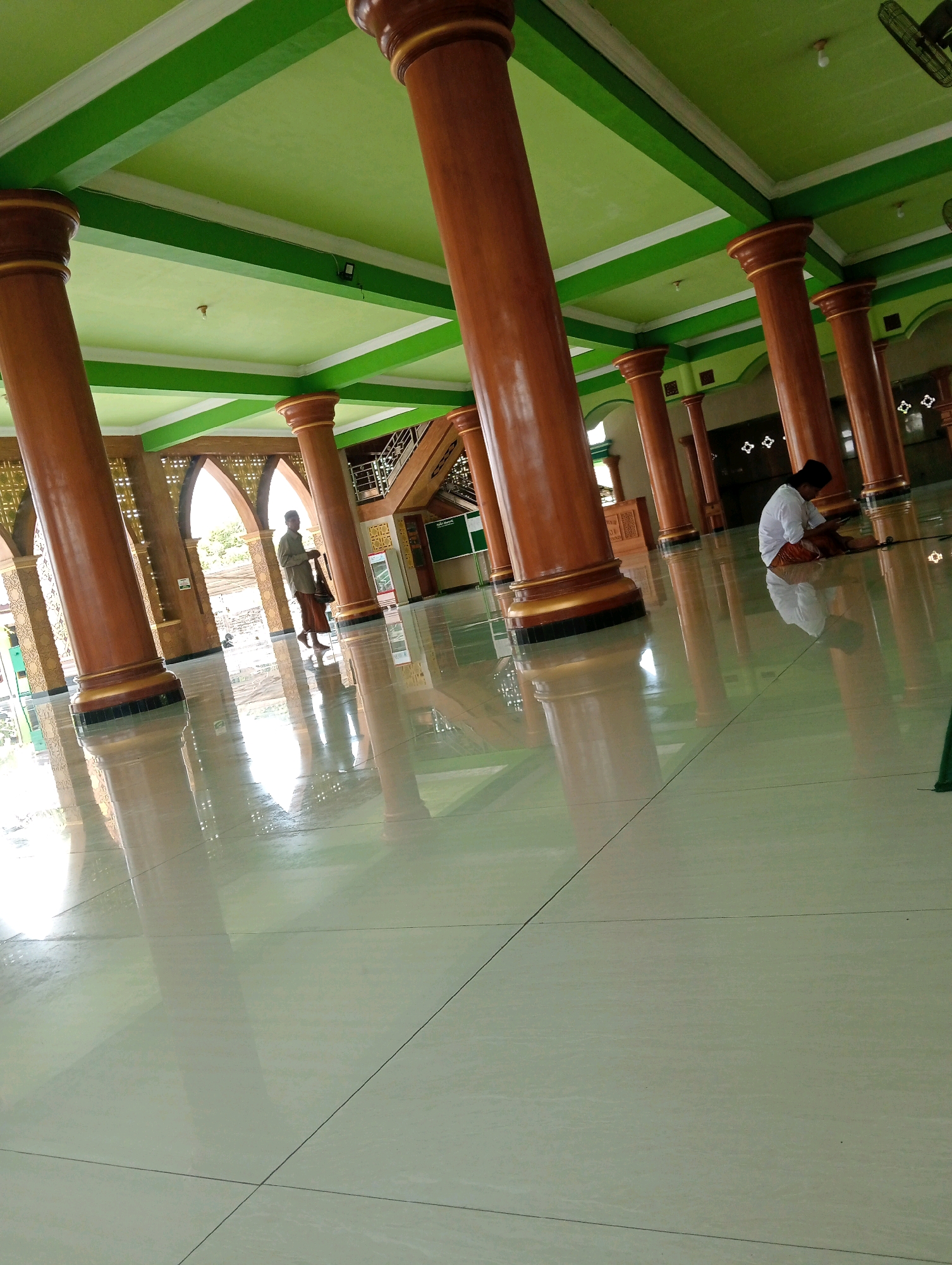In the Soko, Indonesia city, this image captures the interior of a mosque. The scene is dominated by large, polished brown columns supporting a bright green ceiling with white trim. The floor is a highly reflective light-colored tile, mirroring the columns and ceiling, creating a sense of spaciousness. To the right, a person wearing a white shirt and orange-brown trousers is sitting on the floor, facing away from the camera, seemingly engaged with a device or reading material. Further back, towards the left, another individual is standing near an arched opening, dressed in a light-colored shirt and brown sarong, holding a cane. The architecture features ornate arched doorways and decorative elements on the walls. The overall ambiance is serene and communal, indicative of a place of worship.