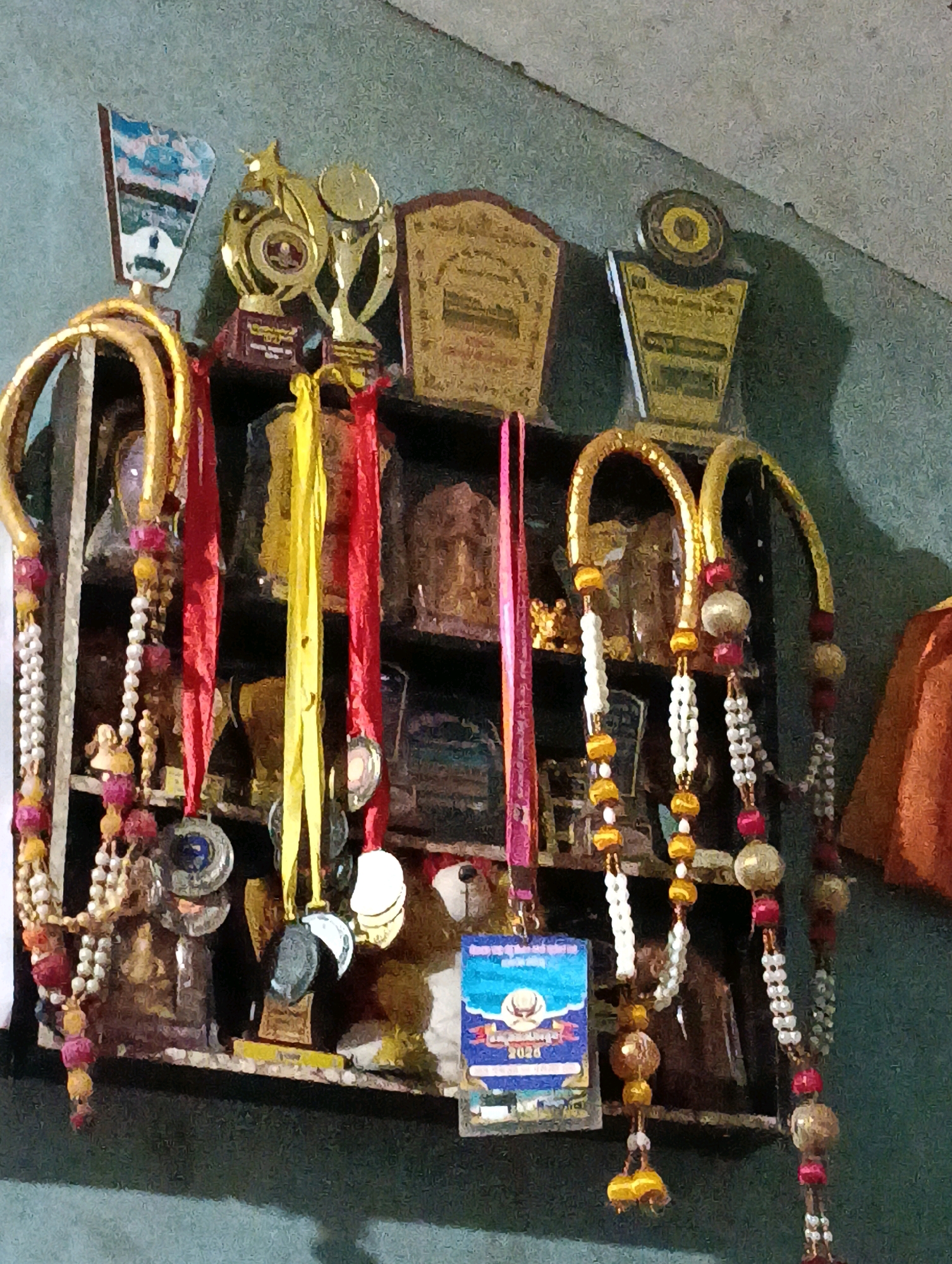 The image depicts a shelf filled with trophies, medals, and decorative beaded strings. The trophies are of various sizes and designs, predominantly gold and bronze in color, with some featuring intricate carvings and emblems. Several medals, some with ribbons, are also visible, hanging from the shelf or attached to the trophies. Decorative beaded strings, composed of small pearls, gold-colored beads, and pink beads, are draped across the shelf, adding a festive touch. A small teddy bear is also present on the shelf, positioned behind some of the trophies and medals. In the background, the wall has a textured, somewhat grimy appearance. The lighting suggests an indoor setting, possibly a display area or a personal collection. The overall impression is one of achievement and commemoration, with a sense of history or personal significance attached to the displayed items. Given the context of the city of Nagore, India, this display could be related to religious events, cultural competitions, or academic achievements within the community.