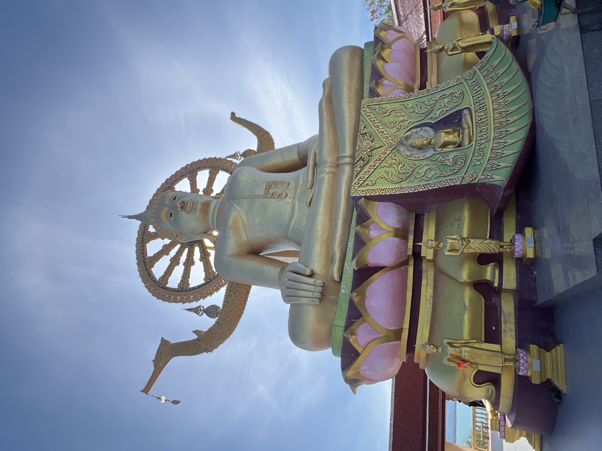 A golden Buddha statue sits in a lotus position with a Dharmacakra above his head in the city of Baan Plai Laem, Thailand. The statue is highly ornamented with details of lotus petals in pink and gold, as well as smaller Buddha statues at the base. The background is a clear blue sky with the sun behind the statue.