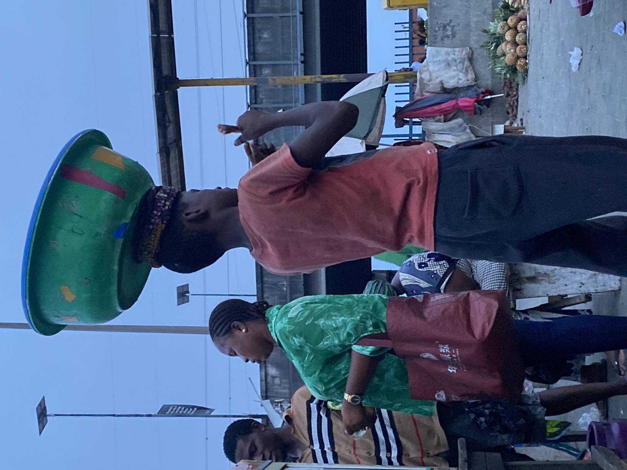 A street scene in Lekki, Nigeria features a woman carrying a large green bowl on her head, balanced with a patterned head wrap. She is wearing a red t-shirt and black pants and making a hand gesture near her face. Beside her is a woman in a green shirt and a brown bag. Further down the frame, a man in a striped shirt is also visible. In the background, there is a bridge-like structure with metal beams, a pile of pineapples on the sidewalk, and other market-related objects. The sky is overcast. The scene appears to be at a busy market location.