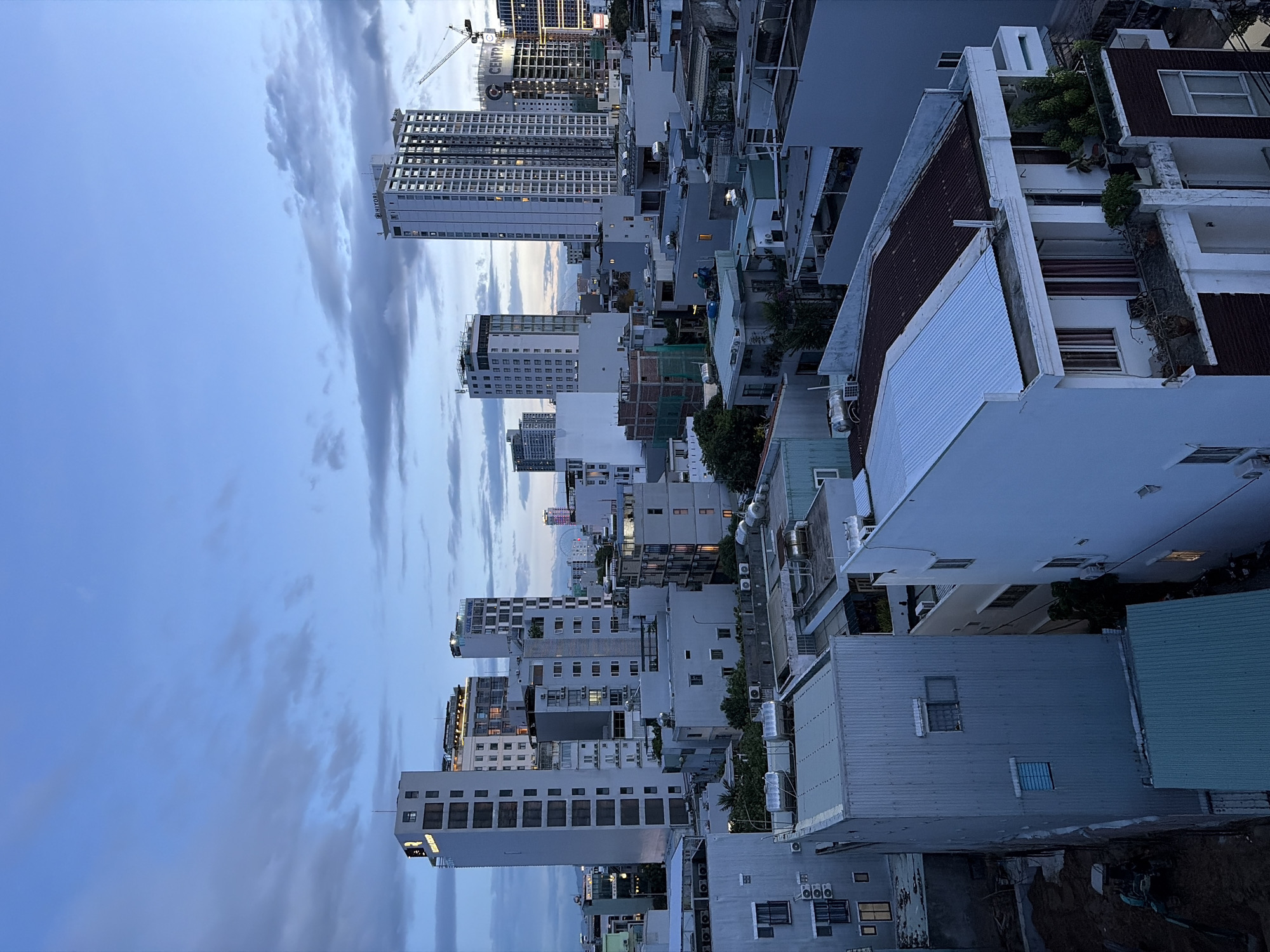 This is a high-angle, full shot of a city skyline in Đà Nẵng, Vietnam, at dusk. The sky is a deep blue, transitioning to lighter hues near the horizon, with scattered gray clouds. The scene is dominated by a dense collection of buildings, ranging in height from low-rise structures to tall, modern skyscrapers.
In the foreground, several lower buildings with corrugated metal roofs and white facades are visible. Some have visible air conditioning units and water heaters on their rooftops. One particularly tall, narrow building on the left has a dark gray metal exterior with small, rectangular windows. Adjacent to it is a lighter colored building with a white facade.
Further back, a cluster of mid-rise and high-rise buildings fills the central and right portions of the image. Some of these buildings have illuminated windows, suggesting occupancy. Notable structures include a tall, white skyscraper with a grid-like pattern of windows on the far right, and a slightly shorter, white building with visible signage that appears to read "CENTRAL". Another prominent skyscraper in the center-left is also white and features a grid of windows. A construction crane is visible behind the "CENTRAL" building, indicating ongoing development.
In the distance, a Ferris wheel can be seen, illuminated against the twilight sky. The overall impression is one of a developing urban environment with a mix of older and newer architecture. No people are visible in the image.