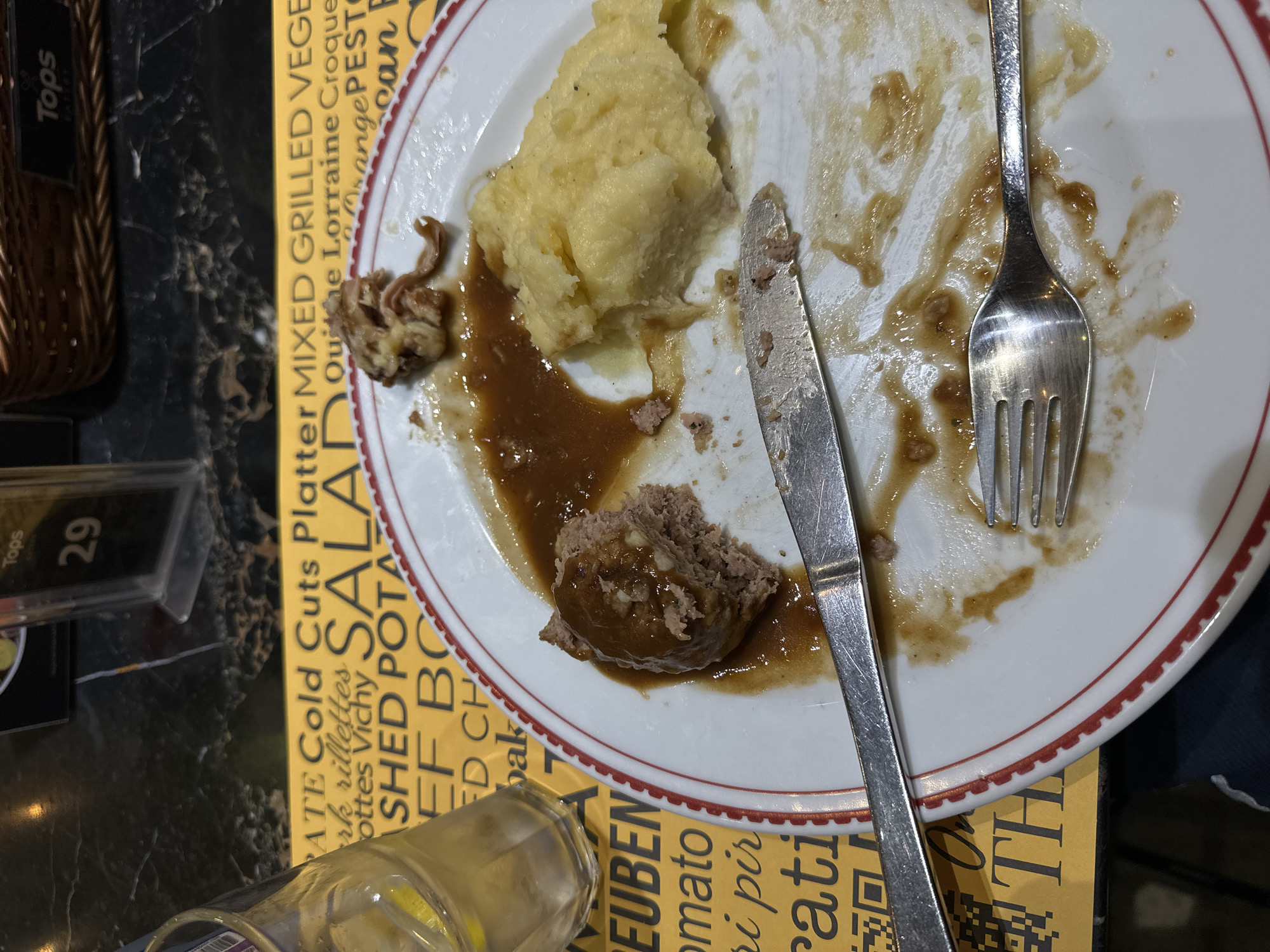 A close-up shot captures a partially eaten meal on a white plate with red trim, set against a yellow and black patterned background. The meal consists of a meatball covered in brown gravy and a scoop of mashed potatoes. A fork and knife are resting on the plate, with remnants of food and gravy scattered around. To the left of the plate, a glass of water with a lemon wedge is visible. The yellow background features text that appears to be a menu, with words like "COLD CUTS," "SALAD," and "POTATO" partially visible. The scene suggests a dining environment, possibly a restaurant. The image does not provide specific details about the location in Wichita, Thailand.