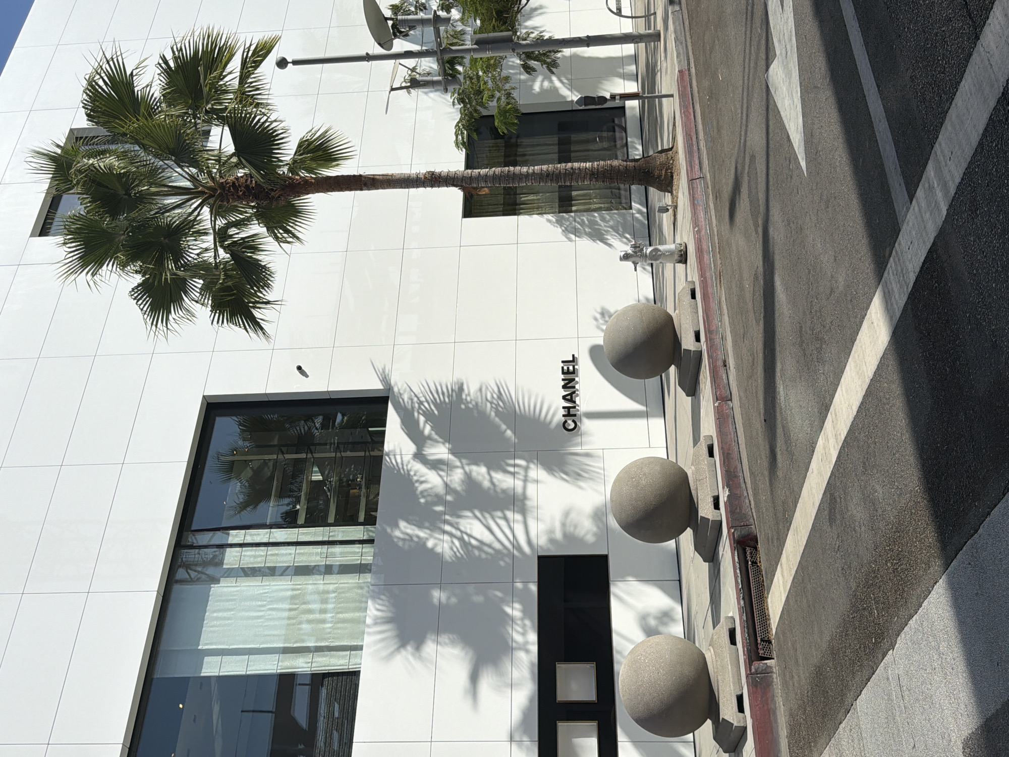 The image depicts the exterior of a modern building with a white facade, featuring a large glass window on the upper left and a prominent "CHANEL" logo on the lower right. In front of the building, there are three large spherical stone planters. A palm tree stands tall to the right of the planters. A sidewalk runs alongside the building, with a street in the foreground. The scene appears to be in Beverly Hills, California, given the architectural style and the presence of palm trees, characteristic of the area. The lighting suggests a bright, sunny day, casting distinct shadows from the palm fronds onto the white building. No people are visible in the image.