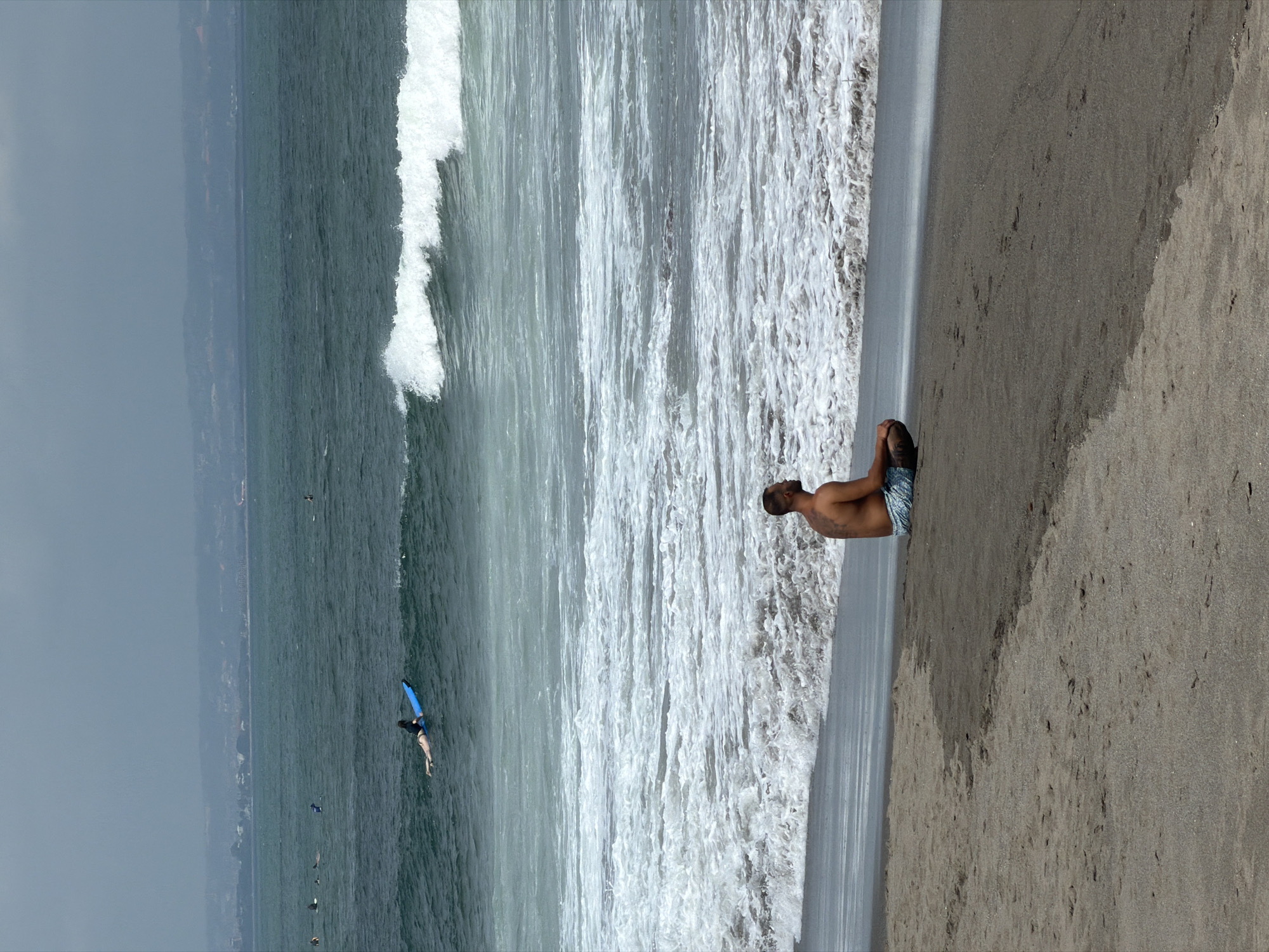 A man with tattoos on his back sits in a meditative pose on the wet sand of a beach in North Kuta, Indonesia. He wears patterned swim trunks. The ocean stretches out before him, with waves crashing on the shore. In the distance, several surfers are visible on the water. The sky is overcast, and a hazy coastline can be seen in the background.
