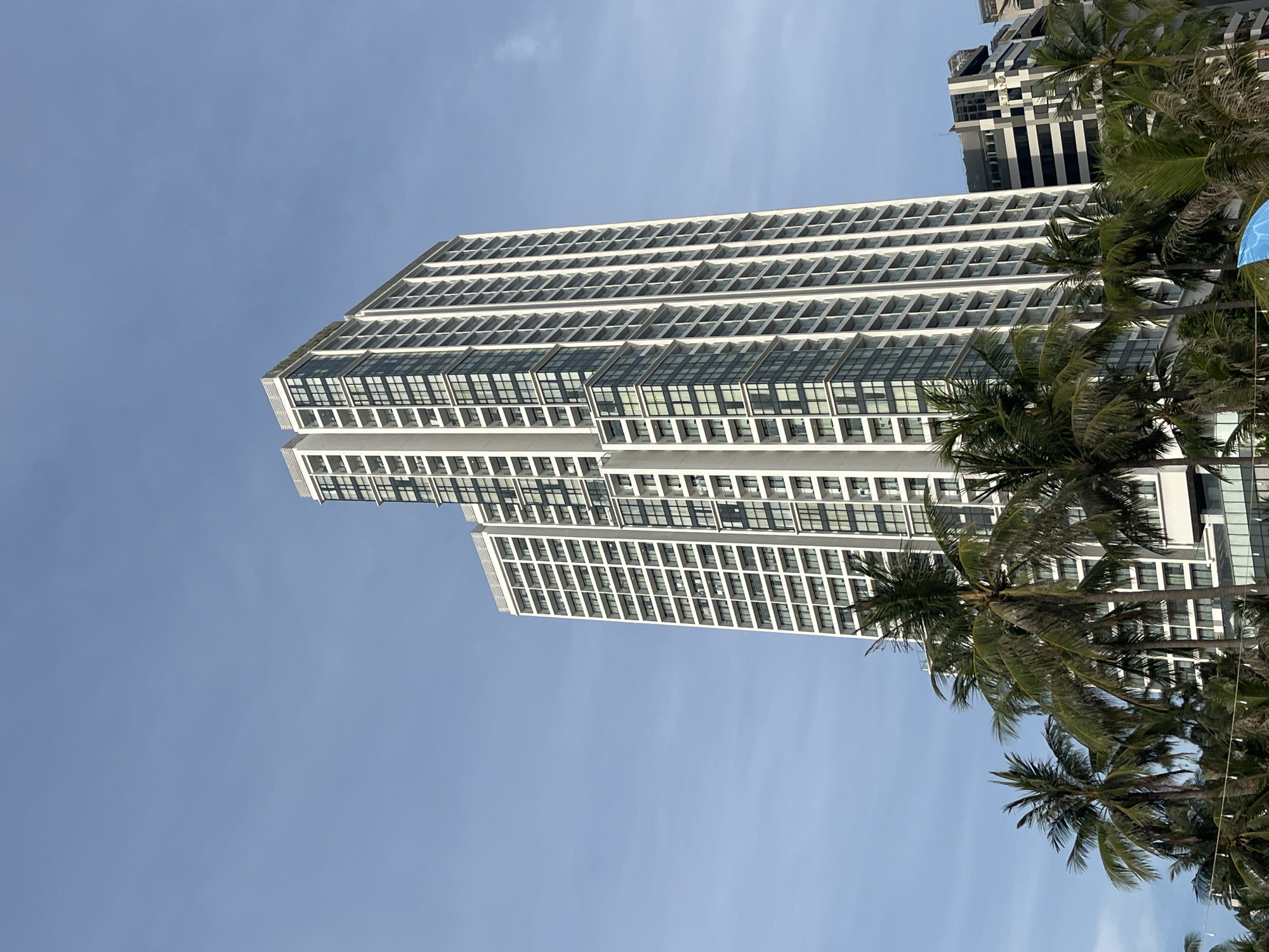 The image shows a tall, modern white building with many windows, likely an apartment complex or hotel, in Nha Trang, Vietnam. Palm trees are visible in the foreground, partially obscuring the lower portion of the building. The sky is clear and blue. The building has a stepped design on the left side and a more uniform facade on the right. Another shorter building with horizontal dark and light stripes is visible to the right of the main building. The scene suggests a tropical urban environment.