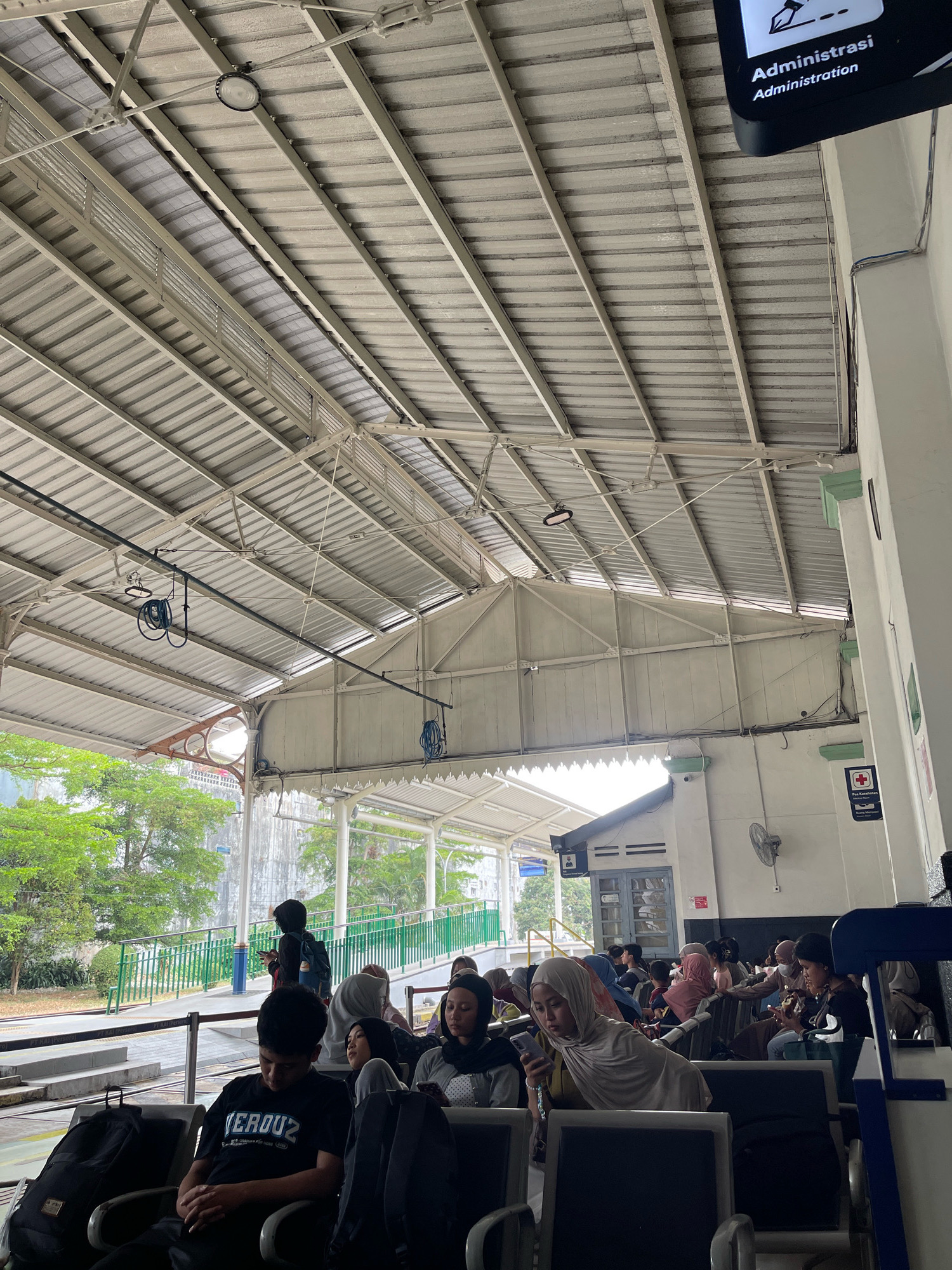 The image captures a waiting area at a train station, likely in Sukabumi, Indonesia, with a vast, corrugated metal roof supported by a complex network of white metal beams and trusses. The ceiling is illuminated by natural light filtering through the sides and several circular light fixtures. In the foreground, several individuals are seated on dark-colored benches, some looking at their phones, others engaged in conversation. Bags are placed on the floor next to the seating.
A green metal railing separates the waiting area from the train tracks visible in the background, where trees and a building with a white facade and blue trim are situated. A sign for "Administrasi" (Administration) is visible at the top right, indicating the station's administrative office. Another sign with a red cross and the text "Pos Kesehatan" (Health Post) is also present. A ceiling fan is mounted on the white wall. The overall scene depicts a typical bustling train station waiting area.