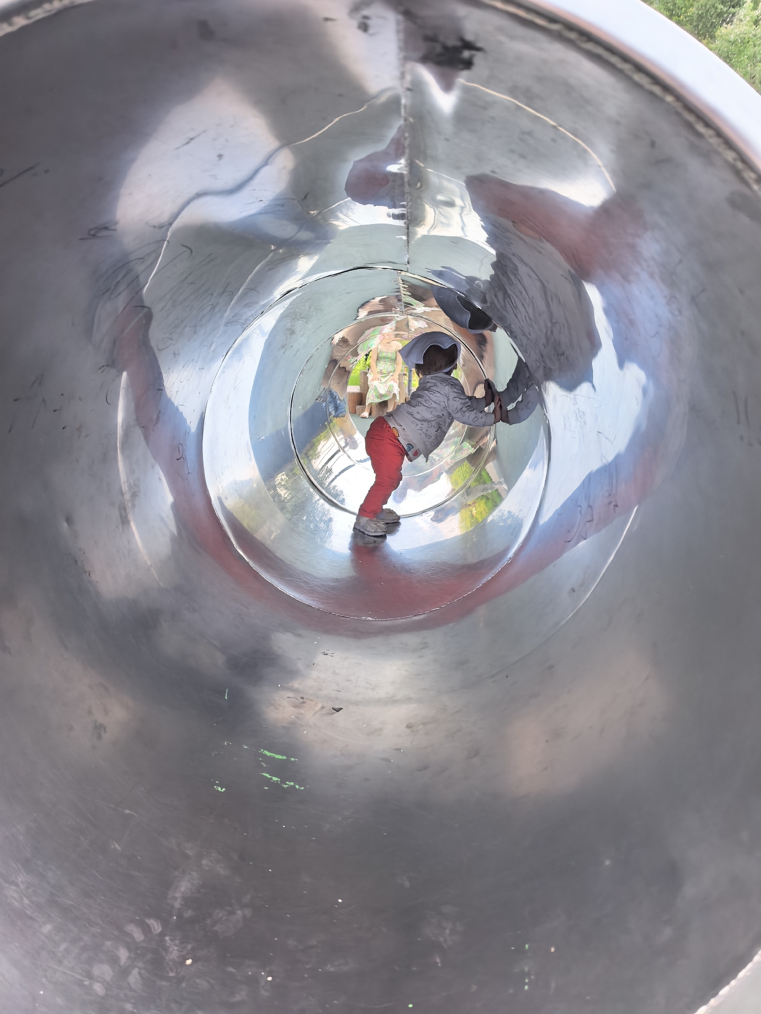 A low-angle, interior shot shows a child with dark hair, wearing a gray sweatshirt with a patterned print, red pants, and white shoes, standing inside a large, reflective, cylindrical metal tunnel, likely a playground slide. The child is facing away from the camera, with their hands pressed against the tunnel wall as if to slow down or stop. Their reflection is visible on the curved, metallic surface. Two other children, one in a green and white dress and another in a blue shirt and black pants, are visible in the distance at the far end of the tunnel, appearing to be at the entrance or exit of the slide. The tunnel walls show scuff marks and some green paint splotches. The environment outside the tunnel, visible through the openings, suggests an outdoor setting with trees and foliage under a bright sky. This scene is located in the city of Sterlitamak, Russia.