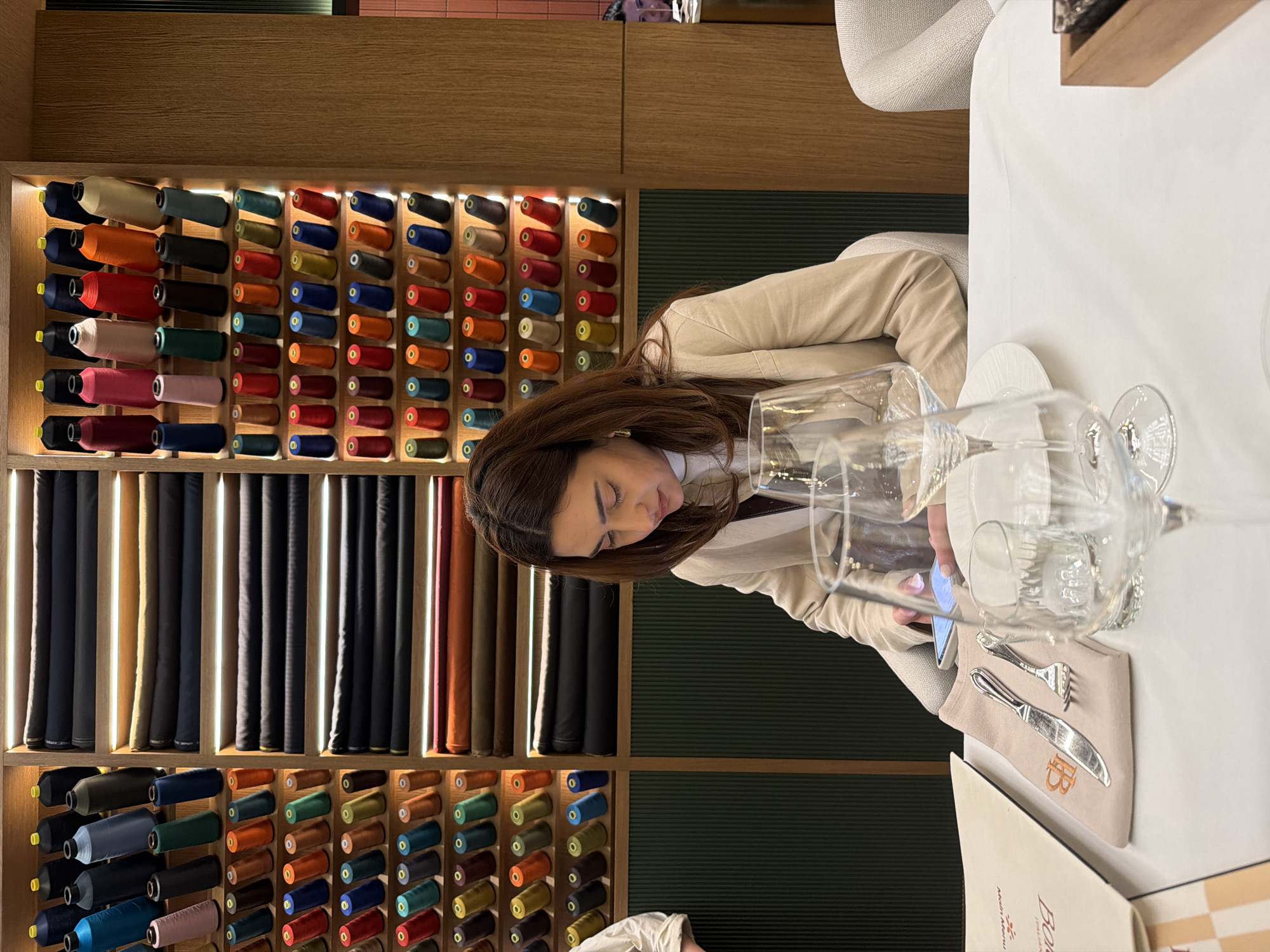 Captured in Yerevan, Armenia, this indoor scene depicts a woman seated at a white-clothed table within what appears to be a bespoke tailoring shop or a fashion-oriented concept store with an integrated dining area. The woman, with long, dark brown hair and dressed in a light-colored jacket, is engrossed in her mobile phone, holding it with her right hand and looking down with a focused expression.
On the table, several clear glass goblets are arranged alongside a beige fabric pouch containing silverware, marked with a logo that includes the letter "B." A menu, partially visible, clearly displays the words "Menu" and "Bor."
The background is dominated by an impressive display of tailoring supplies. Wooden shelves are filled with numerous colorful spools of thread, neatly organized in rows and illuminated by warm lights. Below these, bolts of various fabrics, predominantly in dark shades like black, grey, and brown, are meticulously rolled and stored vertically, interspersed with wooden dividers. The wall behind the shelving features a dark green, ribbed texture. The overall ambiance is refined and organized, suggesting a high-quality establishment where attention to detail is paramount. The visible text includes "Menu," "B," and "Bor" on the table items, and several "0" marks on the thread spools.